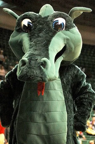 A person dressed in a large dragon costume with big eyes and horns, standing in an indoor setting.