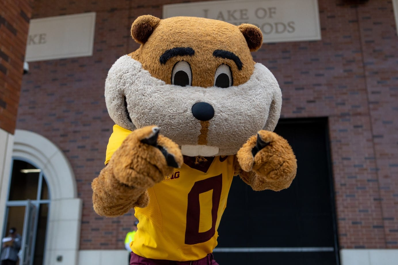 University of Minnesota mascot, goldy Gopher, pointing at the camera, wearing a yellow jersey with maroon letter D, outside a brick building with signs reading 'Make of Foods'