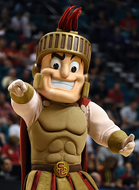 Costumed mascot dressed as a Roman soldier with armor, helmet, and red plume, pointing forward at a sports event.