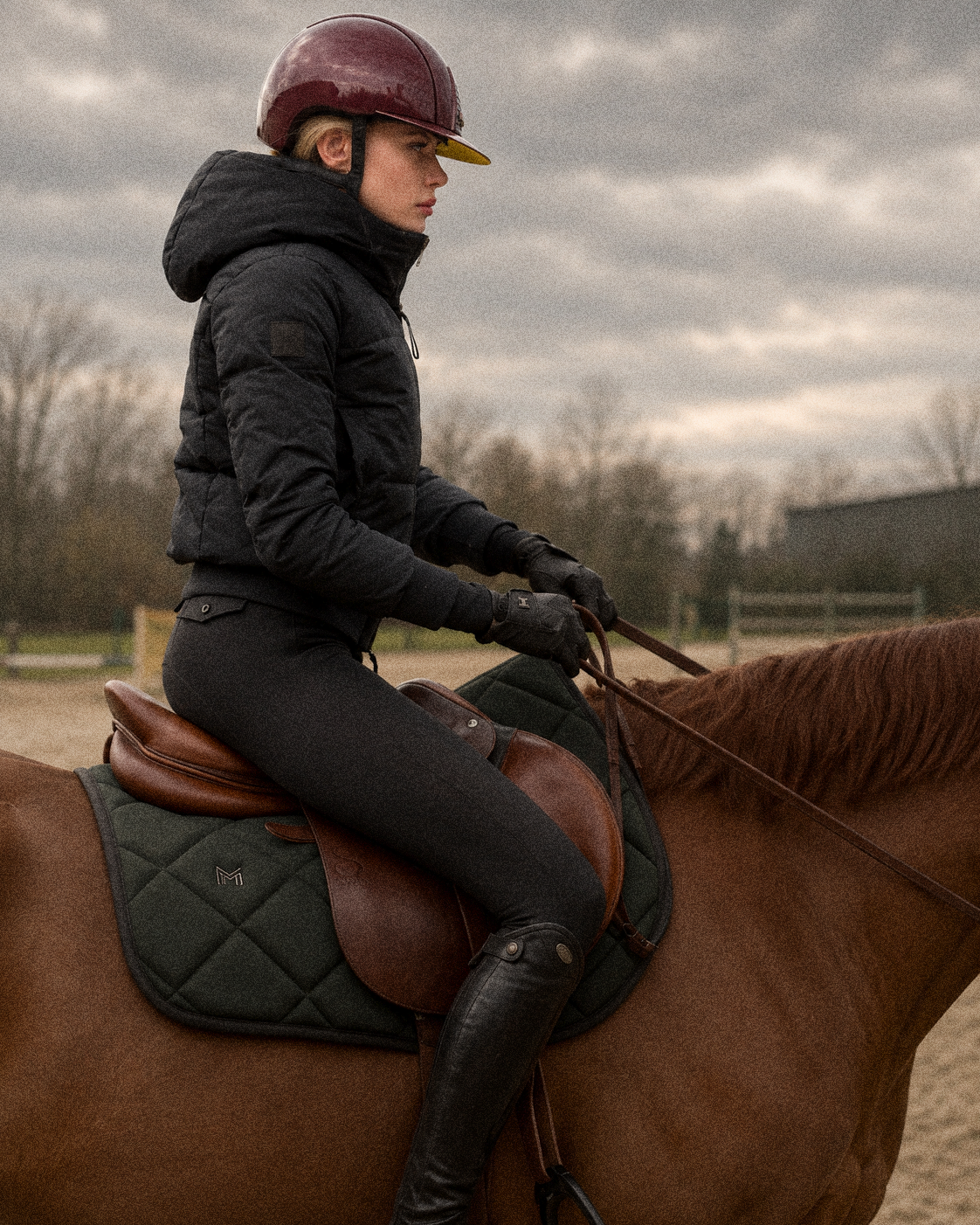 A woman riding a horse outdoors on a cloudy day, wearing riding gear and a maroon helmet.