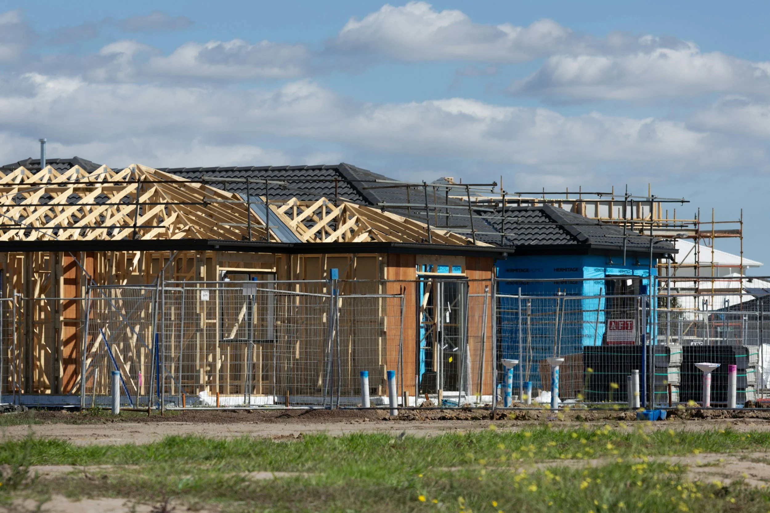 A house under construction with wooden framing, scaffolding, and a partially completed roof with black tiles, set against a cloudy sky.