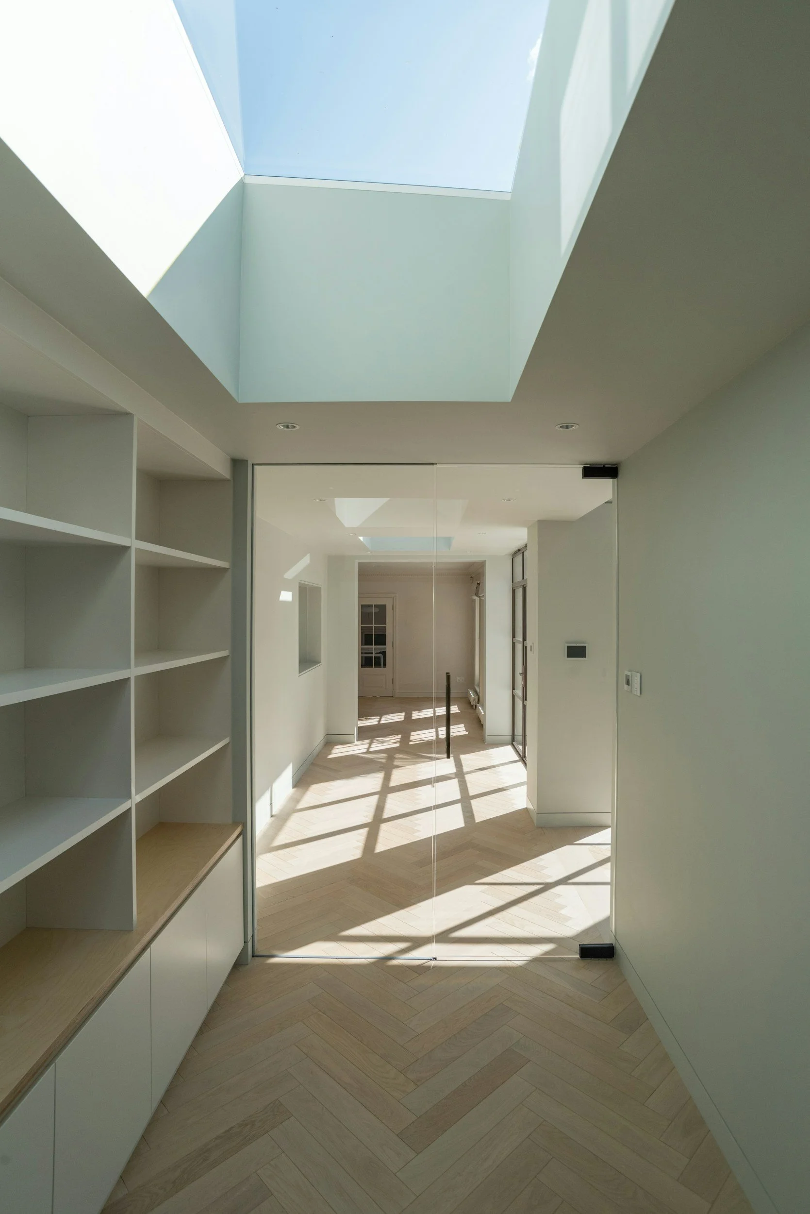 A modern interior hallway with light-colored wooden herringbone flooring, white walls, built-in shelves on the left, and a large glass door on the right reflecting the sunny corridor and blue sky.