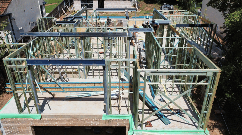 Construction site with a wooden framing structure in progress, including metal beams and workers, on top of a brick building foundation.