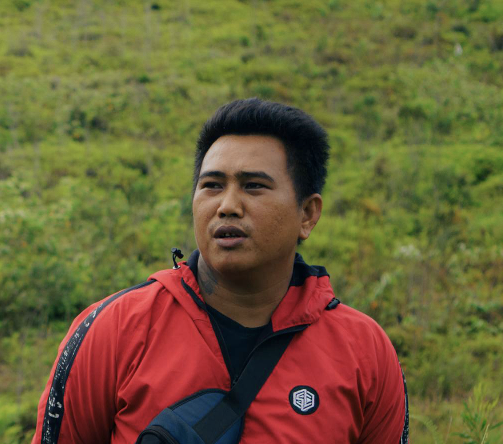 A man wearing a red jacket standing outdoors with green hills in the background.