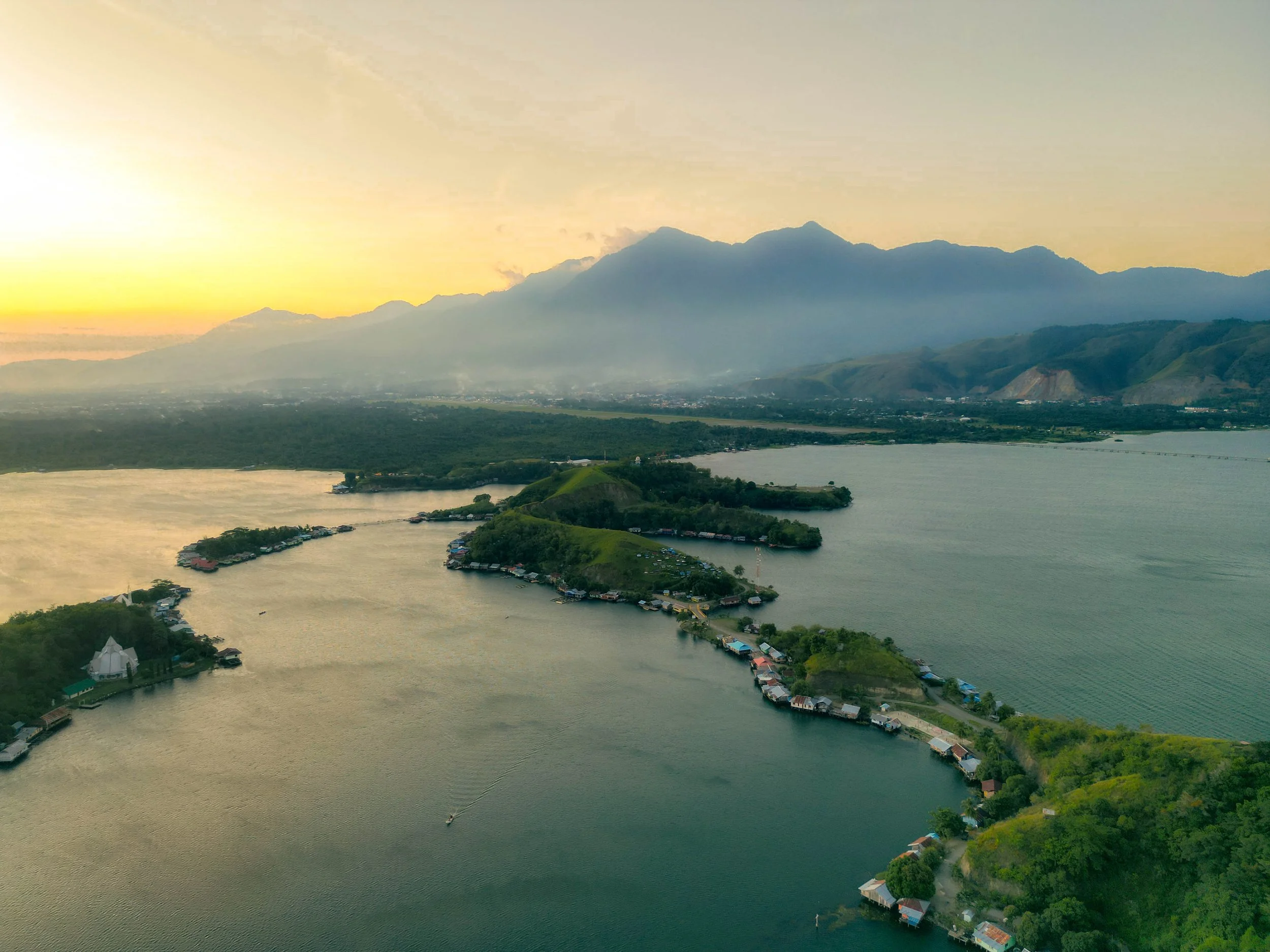 Aerial view of a river with small houses and buildings along the shoreline, surrounded by green hills and mountains during sunset