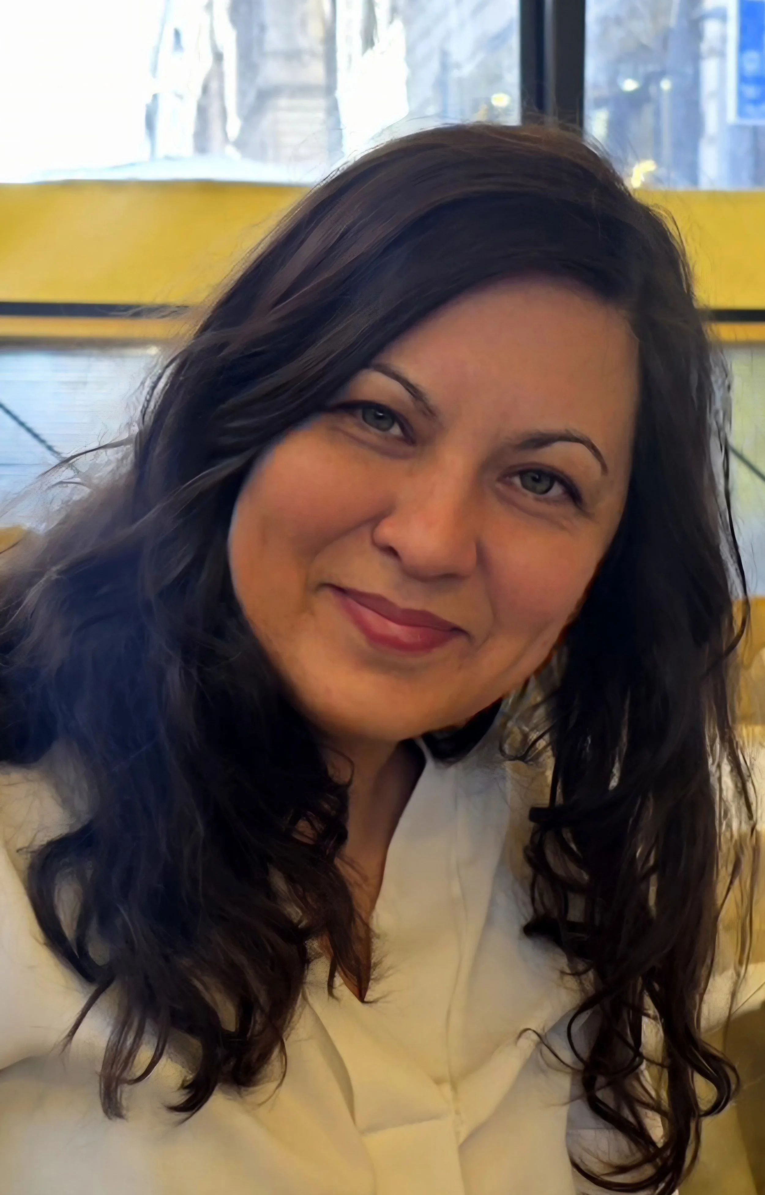 Close-up of a woman with long, dark curly hair, light skin, and blue eyes, smiling, sitting in a yellow booth with a glass window behind her showing a city street scene.