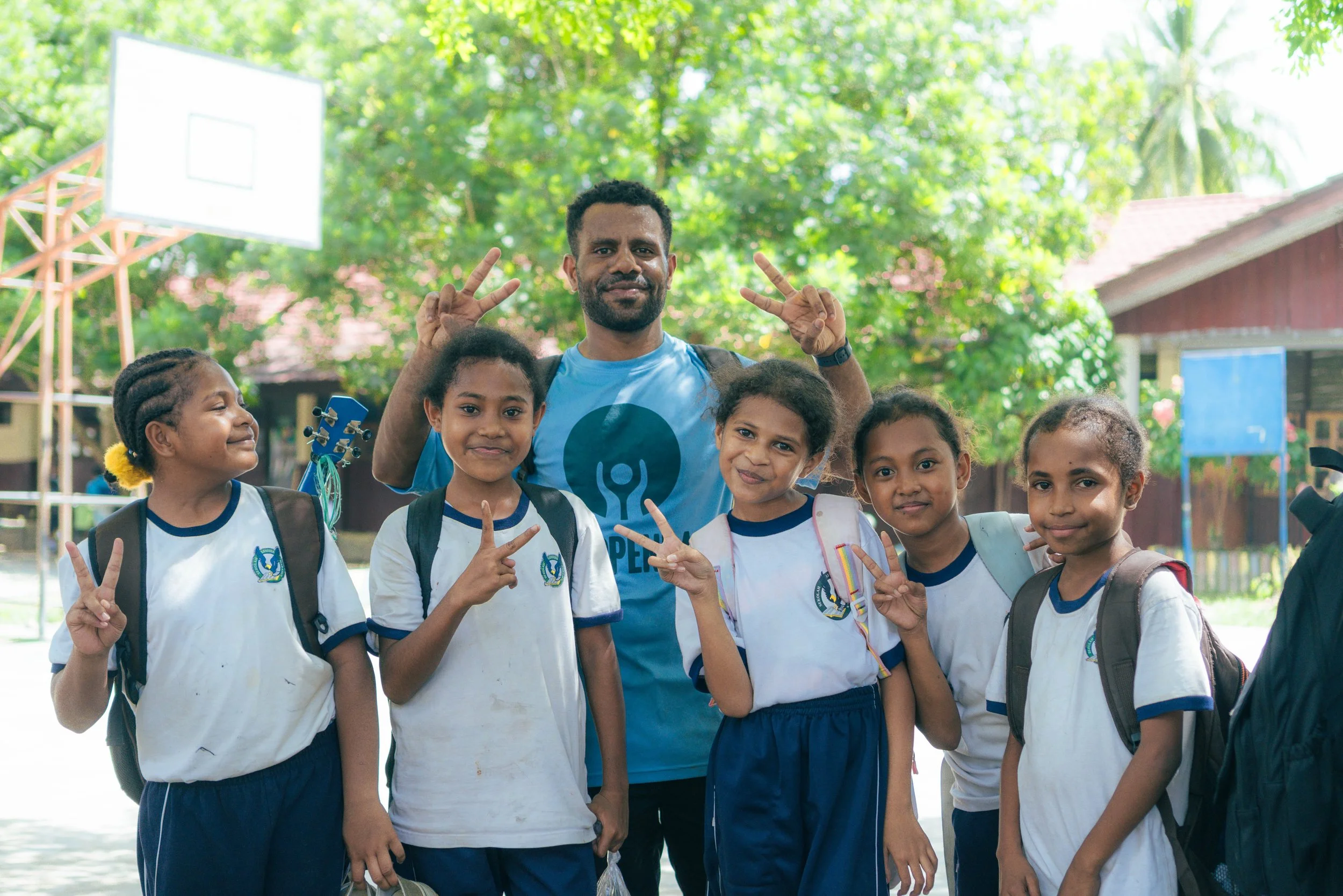 A group of five diverse young girls and one adult man, all smiling and making peace signs with their fingers. They are outdoors on a sunny day with green trees and a basketball hoop in the background, and they appear to be at a school or park.