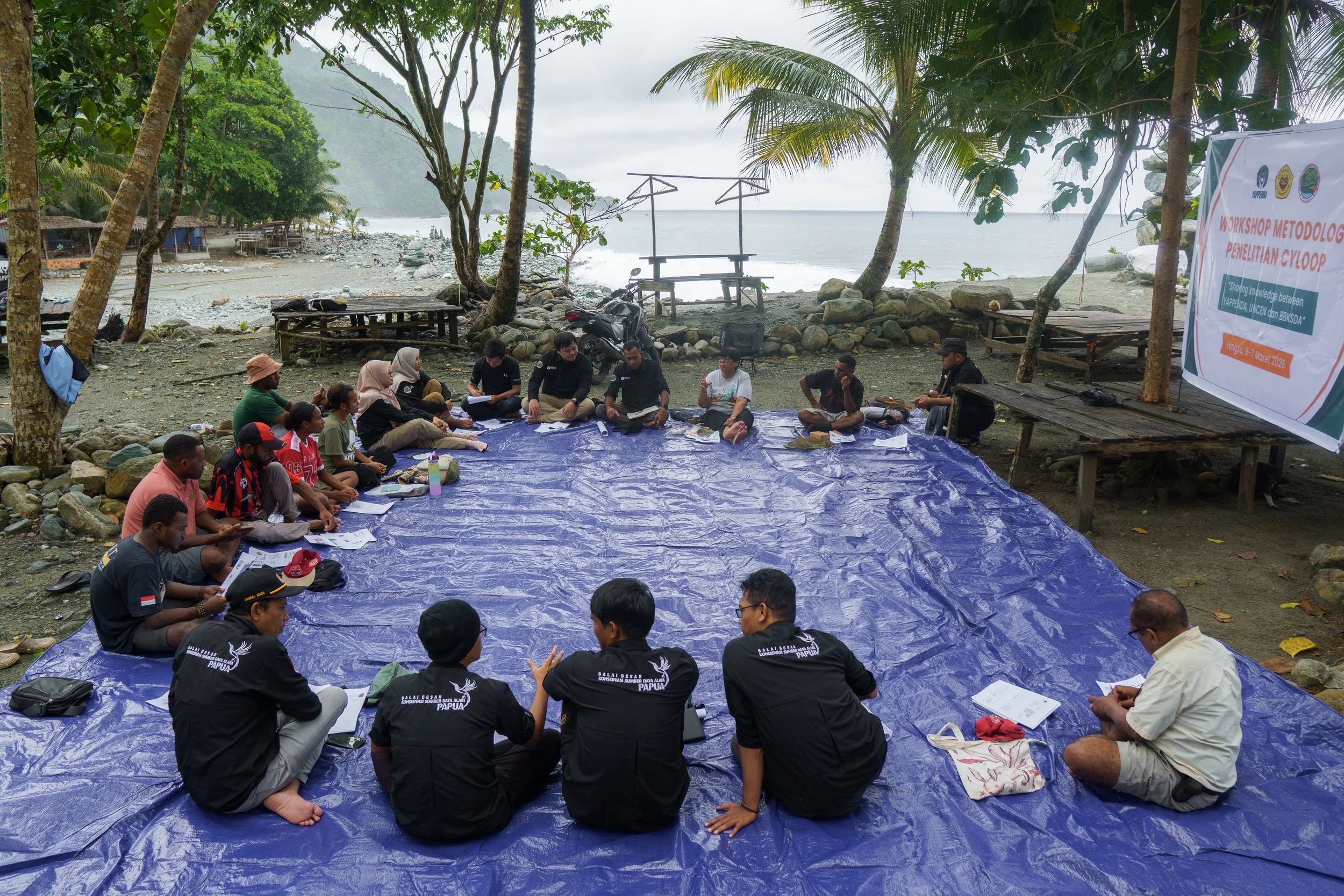 A group of people sitting on a large blue tarp on a rocky beach, participating in a workshop near the ocean with trees and a sign in the background.