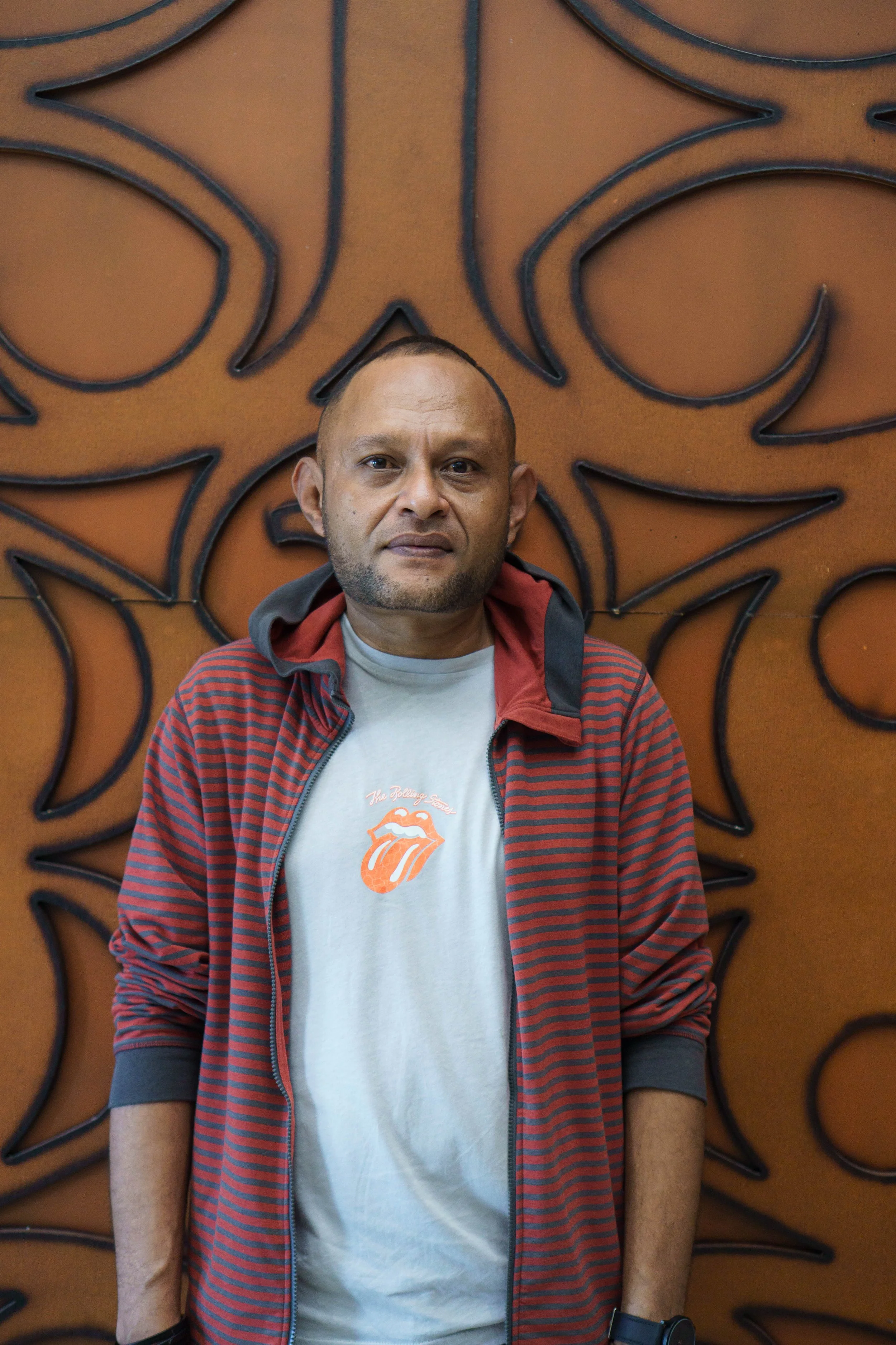 A man wearing a gray T-shirt with a Rolling Stones tongue logo, a red and black striped jacket, and a black wristwatch, standing in front of a decorative, ornate brown wall.
