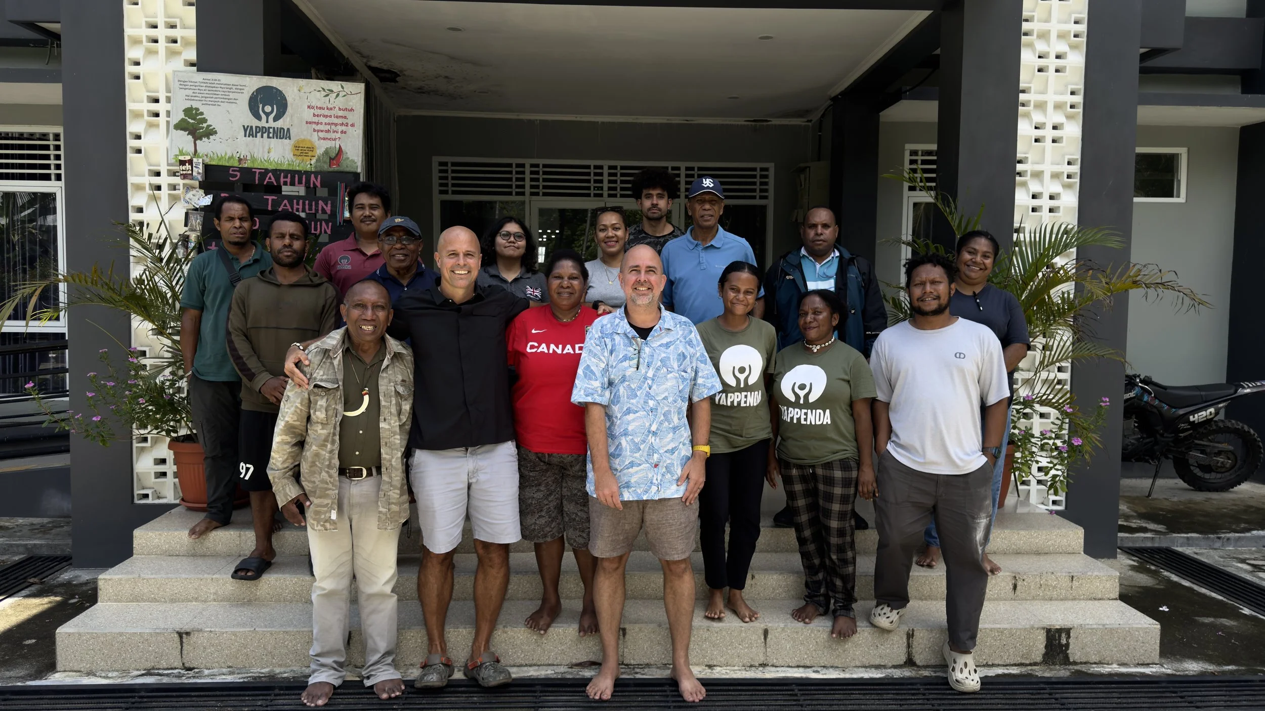 A group of diverse people standing on steps in front of a building with a sign that reads "YAPPENDA." Some are smiling, and a few are barefoot. There are potted plants on either side of the group, and a bicycle is visible on the right side.