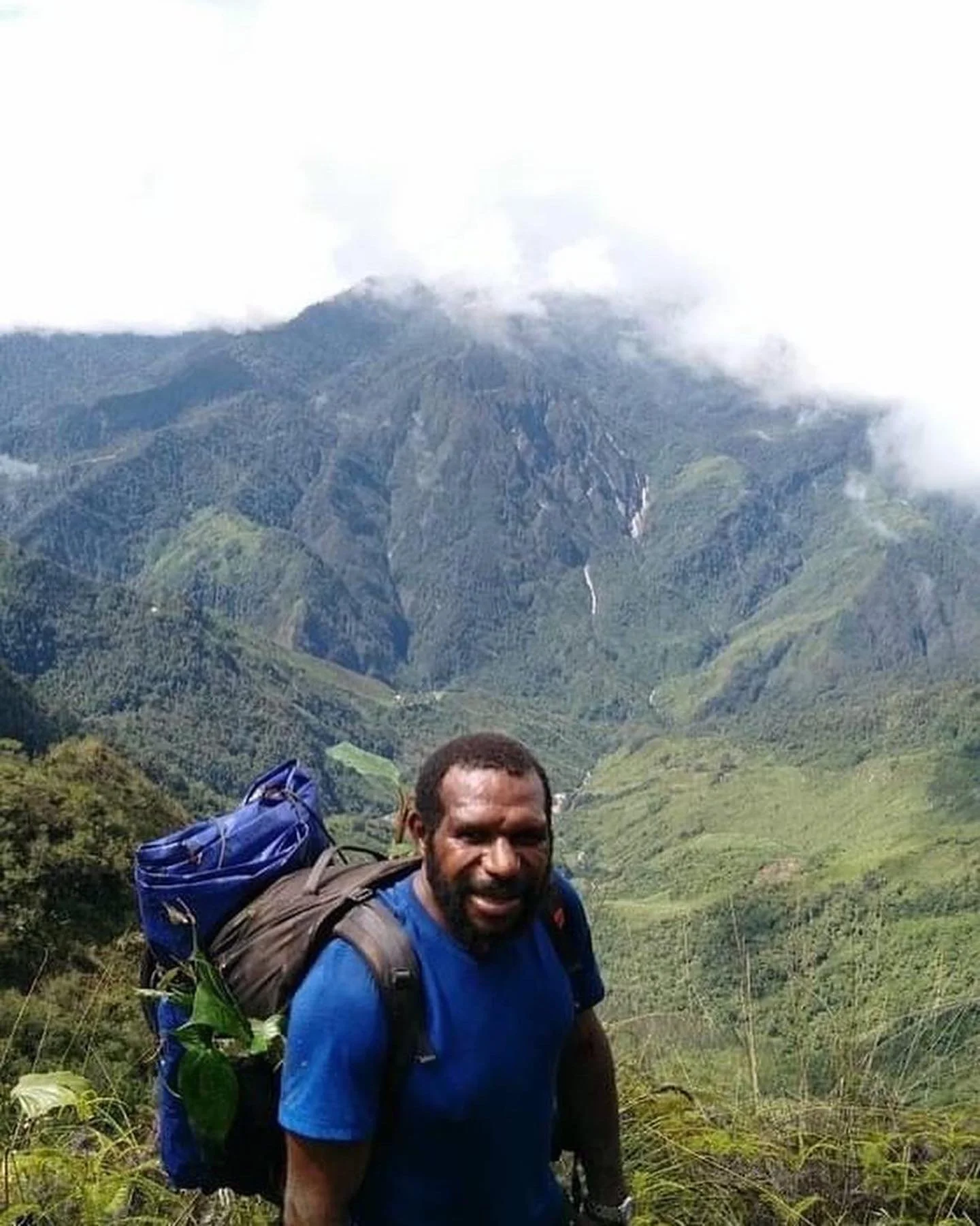 Man with backpack hiking in a lush green valley with a mountain and cloud-covered volcano in the background.