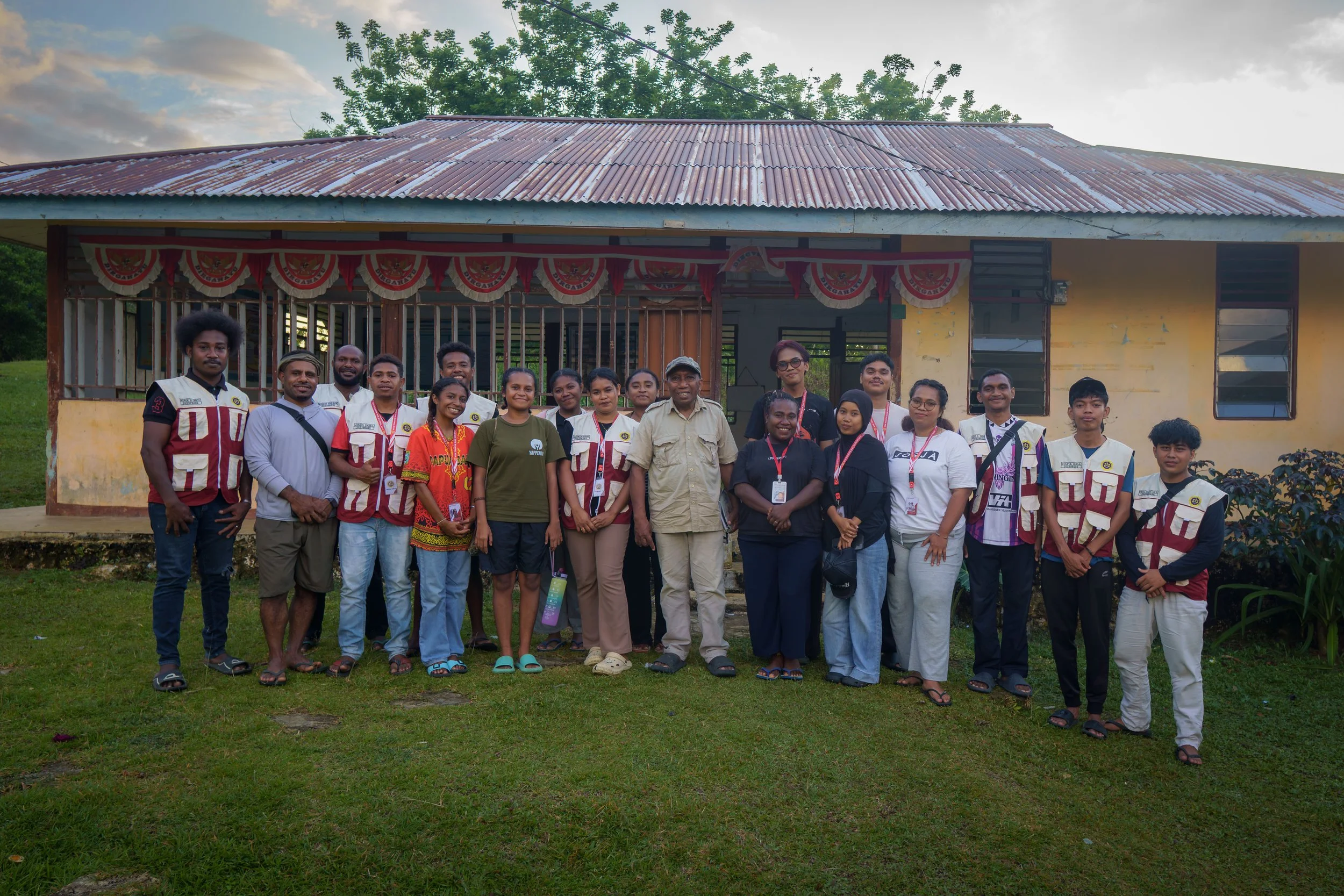 Group of people standing in front of a yellow building with a rusty metal roof, some wearing lanyards, outdoors during daytime.