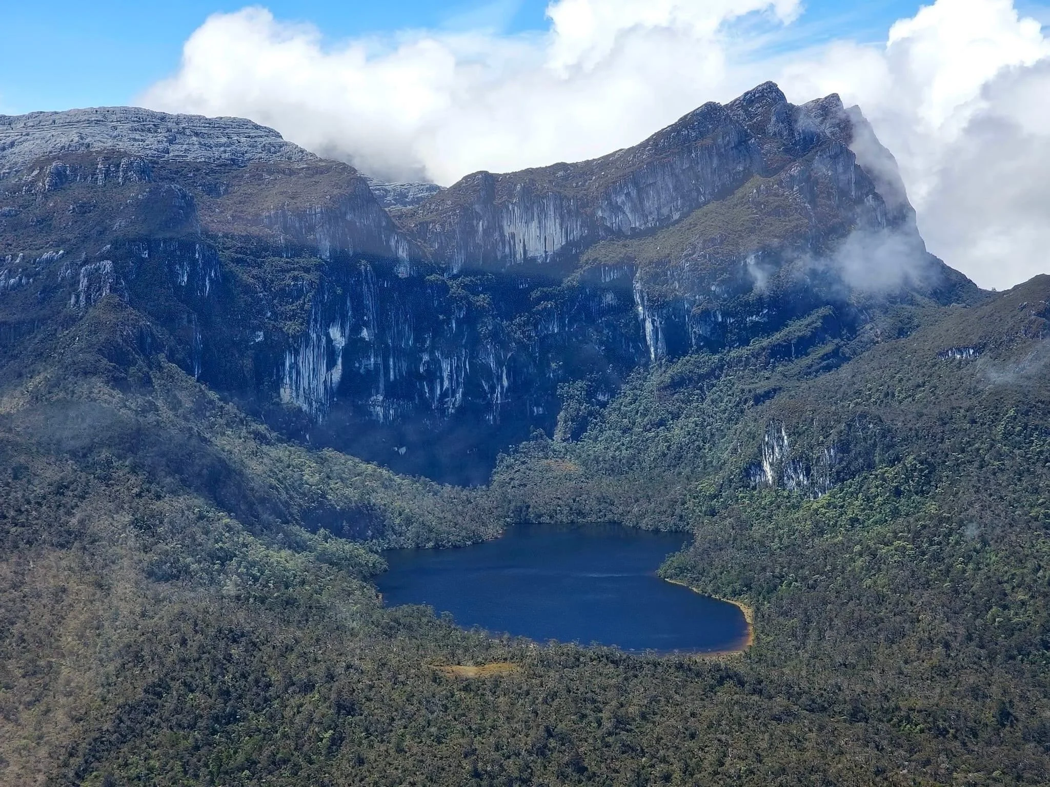 Mountain landscape with rugged peaks partially covered in clouds, dense green forest at the base, and a body of water in the foreground.