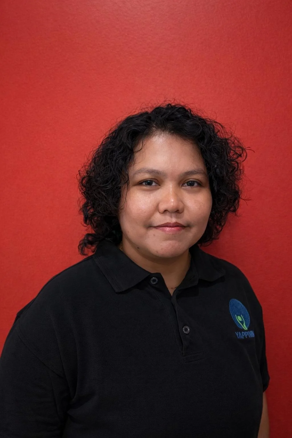 A woman with curly black hair wearing a black polo shirt with a logo, standing in front of a red wall.