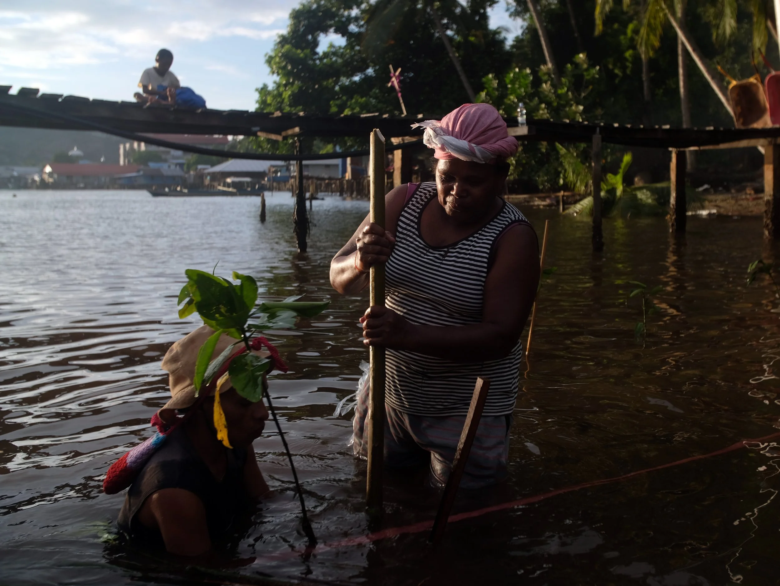 Three women are engaging in a water activity near a waterfront with tropical trees and houses in the background. Two women are in the water holding a stick, and one woman, standing on a platform, is holding a stick or pole, possibly involved in a traditional activity or fishing. The women are dressed in casual clothing and head wraps, with one wearing a striped top.