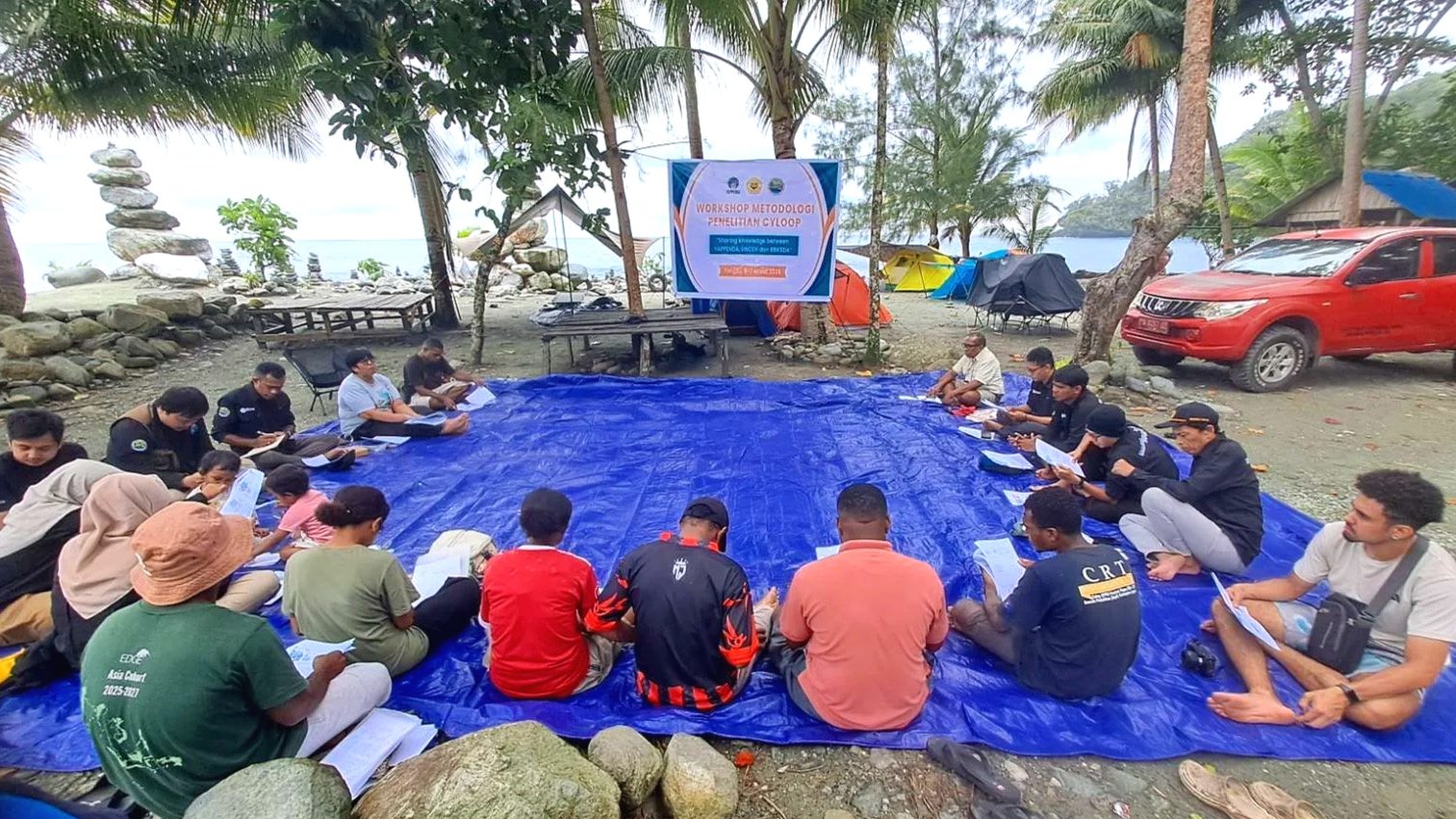Group of people sitting in a circle on a blue tarp outdoors near a beach, attending a workshop with a banner that reads 'Workshop Metodologi Penelitian Cyloop,' surrounded by trees, rocks, tents, and a red vehicle.