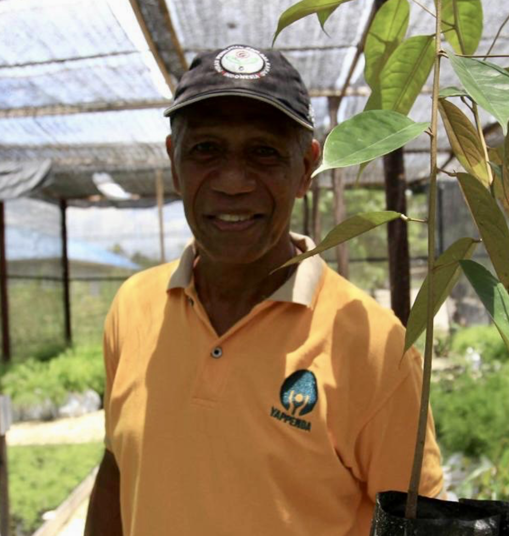 A man with dark skin smiles at the camera inside a greenhouse, wearing a dark cap with a circular logo and an orange polo shirt with a blue and yellow logo on the chest. There are green leaves and plants around him.
