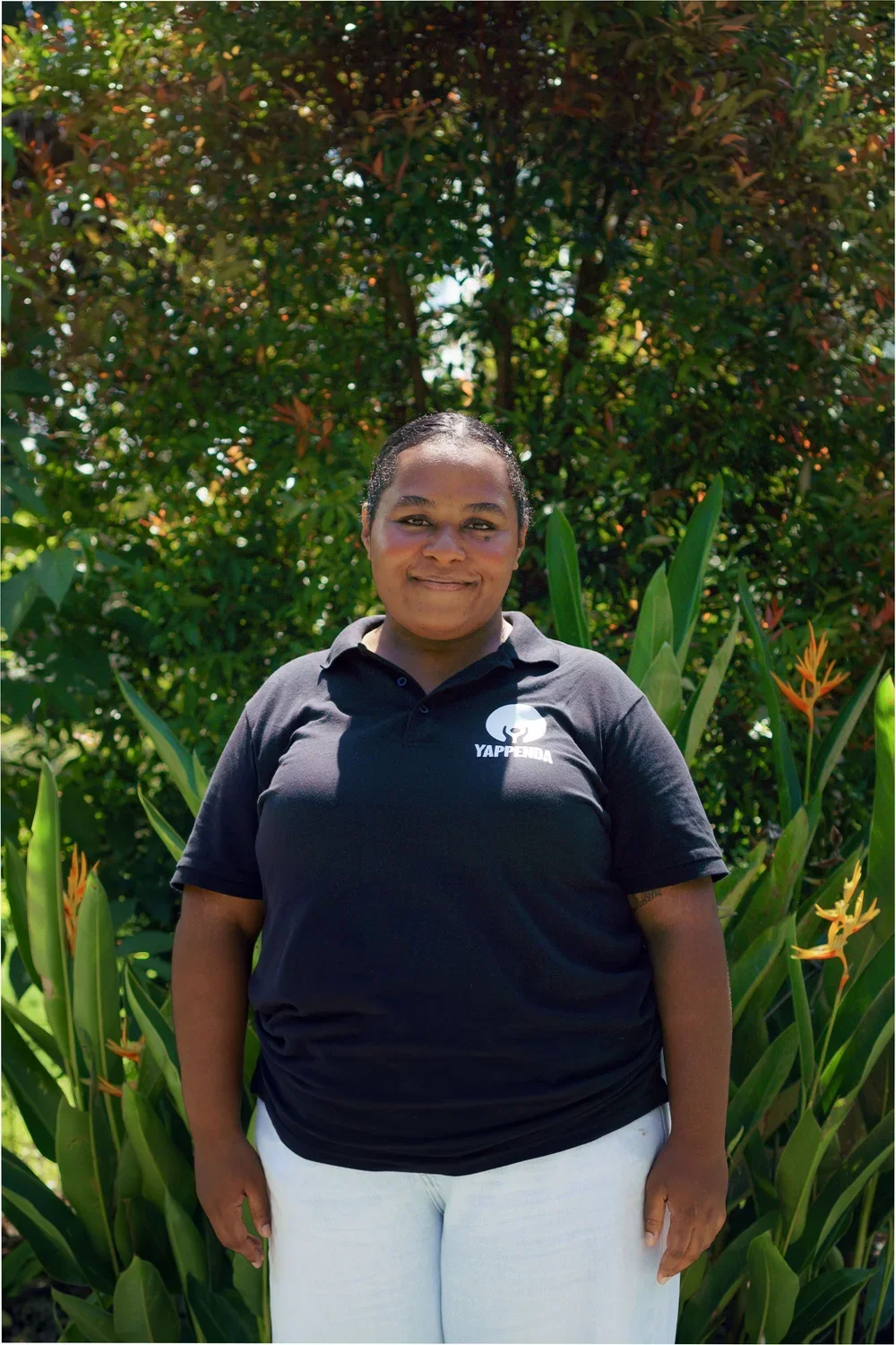 A woman standing outdoors in front of greenery, wearing a black polo shirt with a logo and the word 'YAPPIEDA', smiling at the camera.