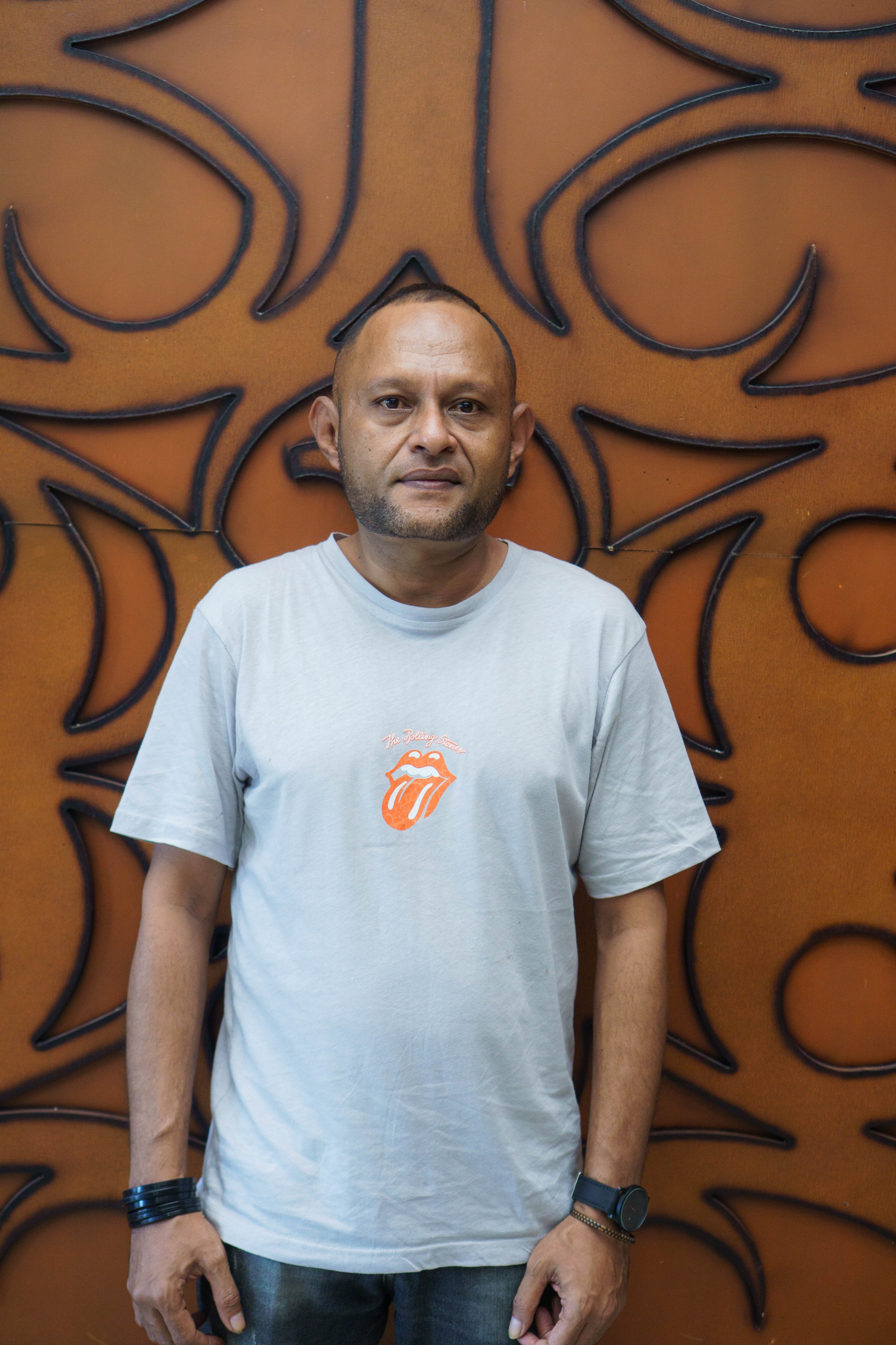 A man standing in front of a decorative orange wall with a black abstract design. He is wearing a light gray T-shirt with a tongue logo and text, along with a black wristwatch and bracelet on his left wrist.