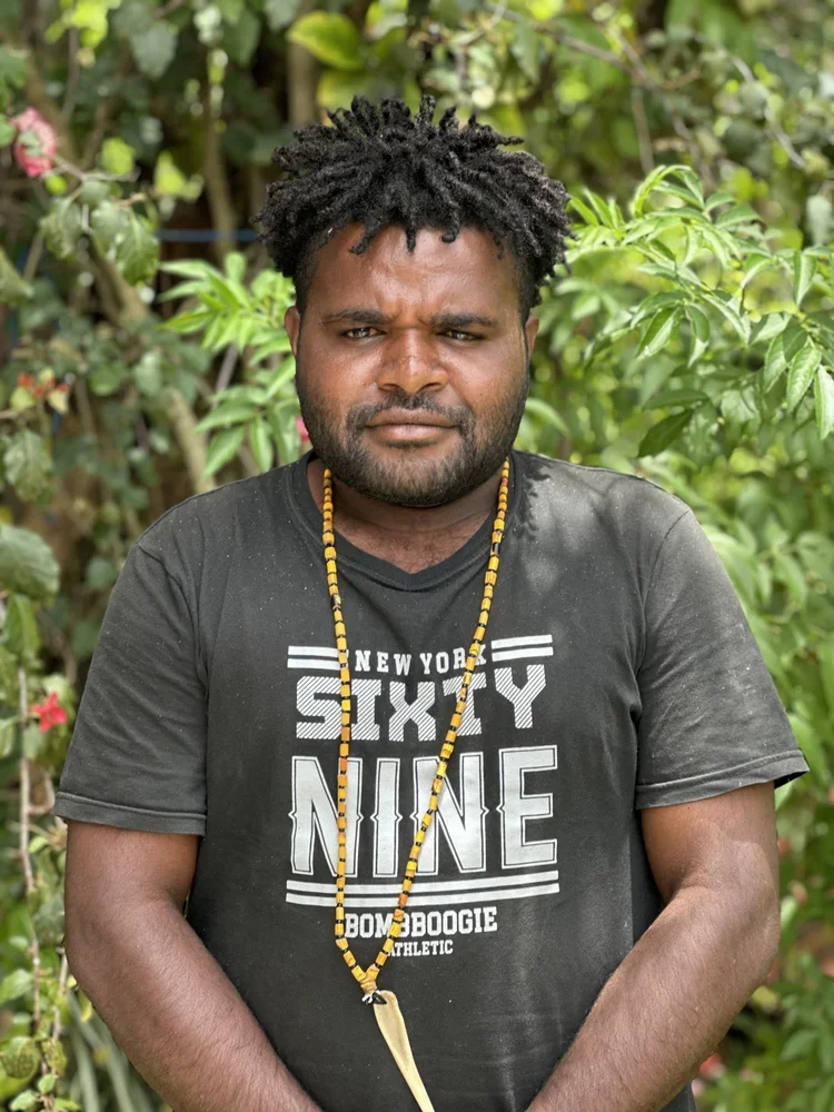 A man with dark, curly hair and a beard standing outdoors amid green foliage, wearing a black t-shirt with white text and a yellow beaded necklace.