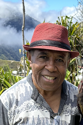 A smiling man wearing a red fedora hat with a black band, against a tropical background with green plants, mountains, and a partly cloudy sky.
