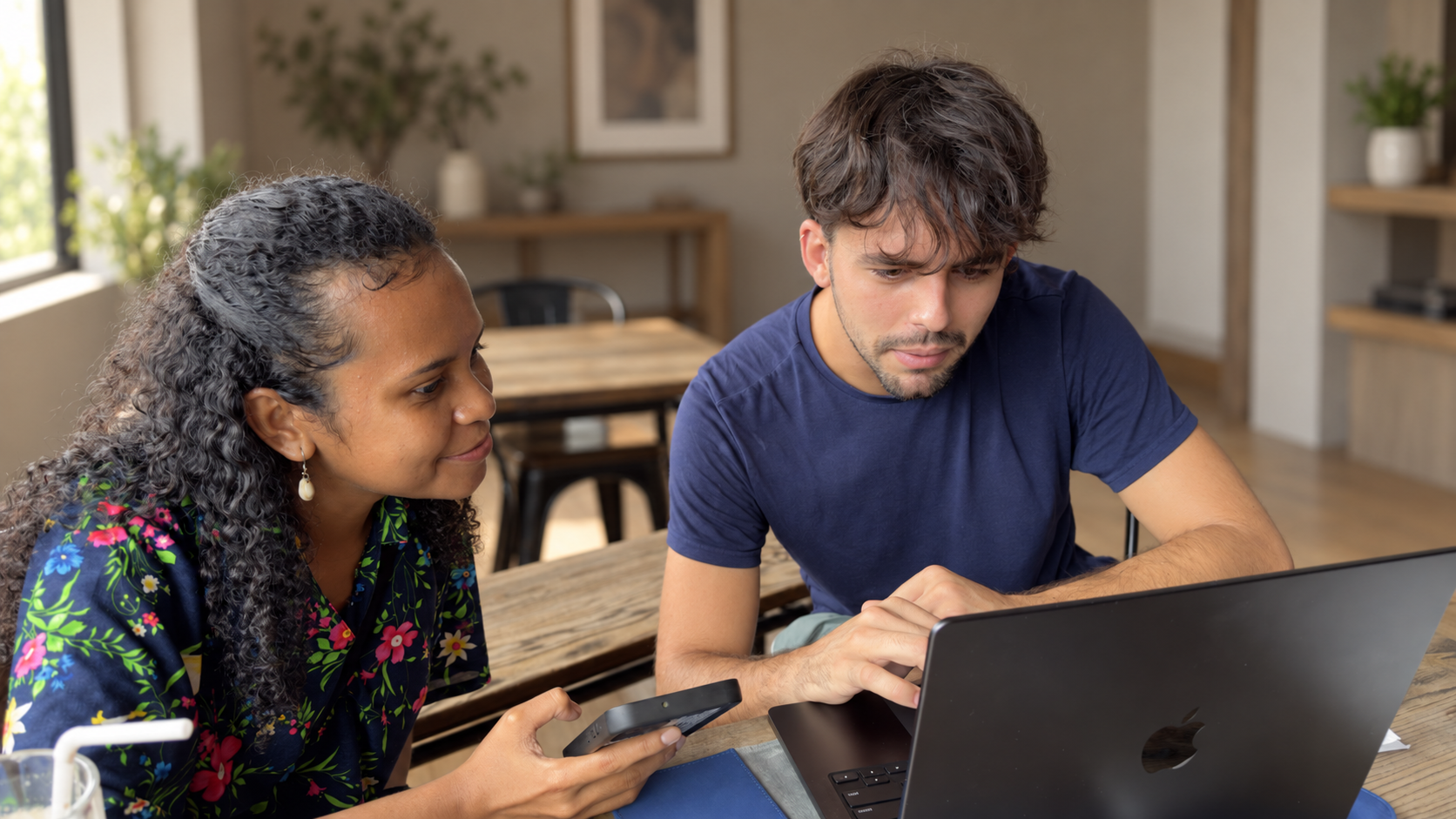 A man and a woman sitting at a wooden table, looking at a laptop and a smartphone in a bright, modern room.