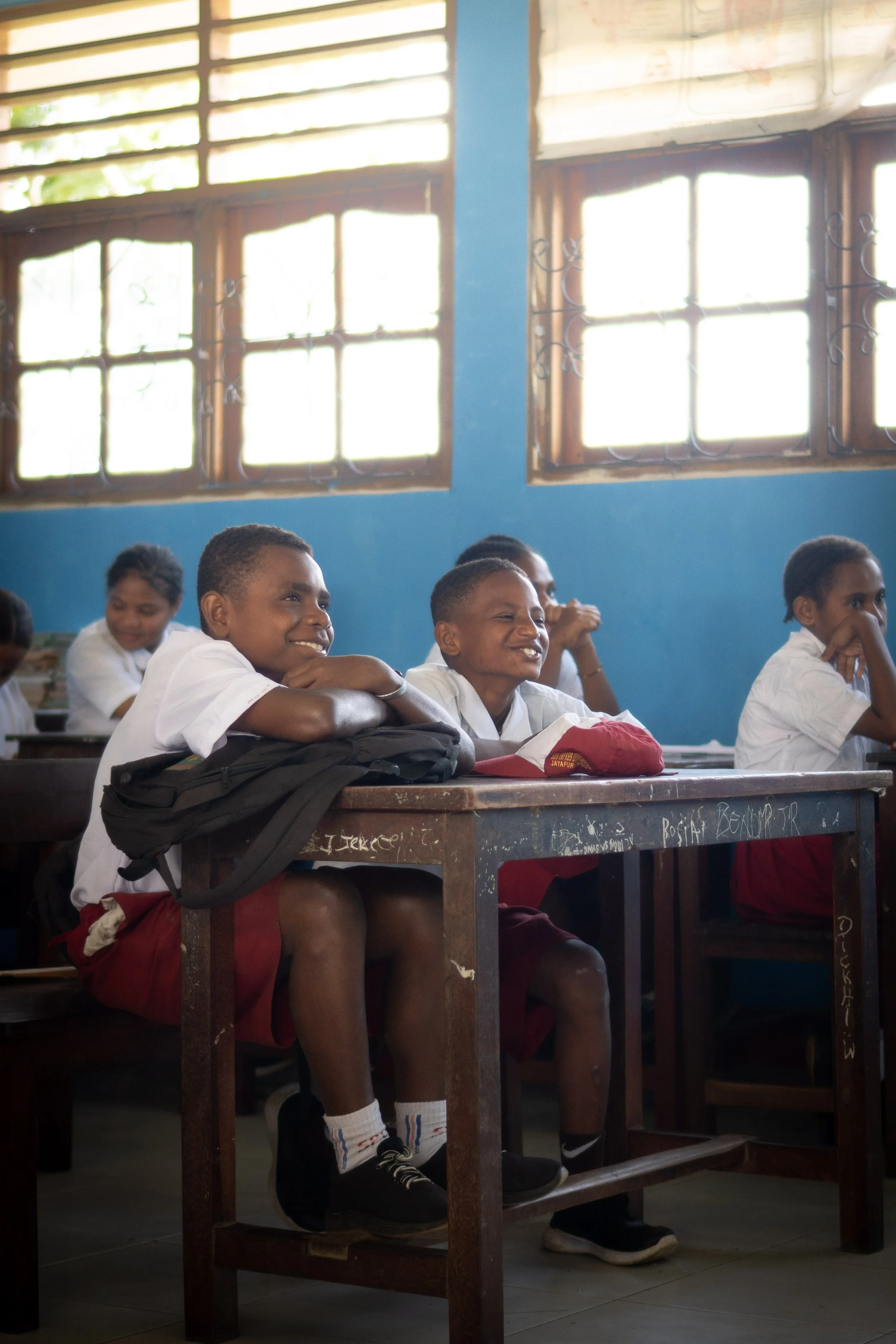 A classroom with students sitting at desks, smiling and paying attention. There are large windows on the blue wall providing natural light.