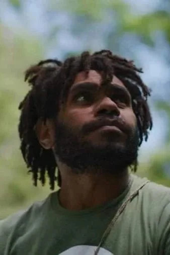 A man with dreadlocks and a beard outdoors, looking up.