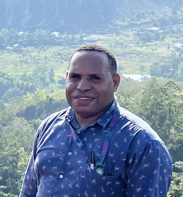 Man smiling outdoors with a green, hilly landscape in the background.