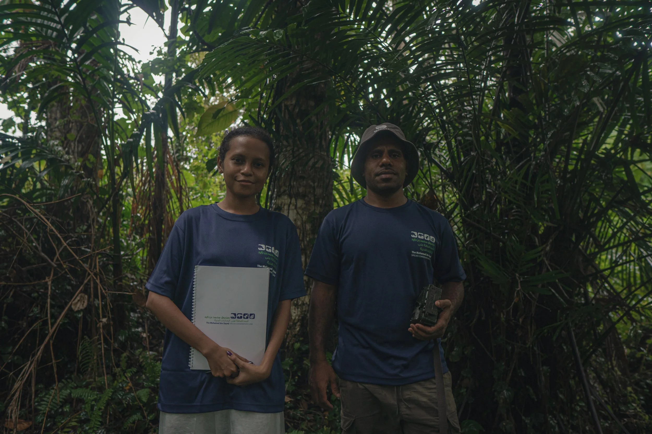 Two people standing in a dense jungle, both wearing navy blue shirts. The woman on the left holds a white notebook, and the man on the right holds a device. They look at the camera with serious expressions.