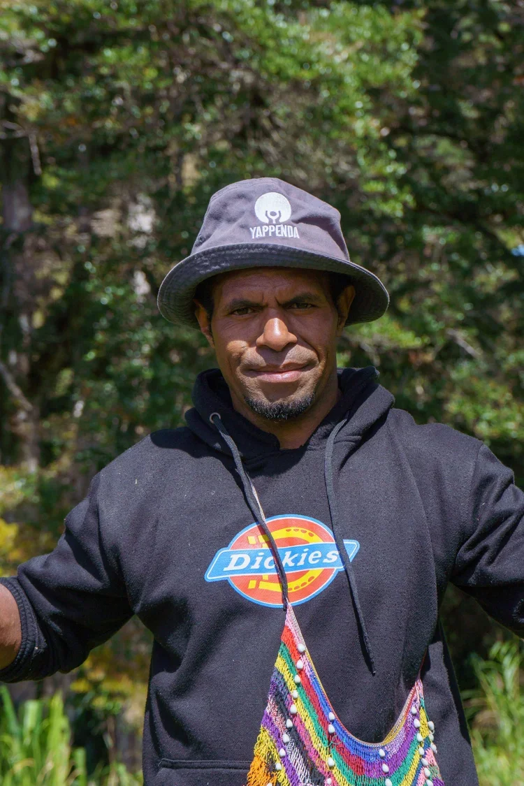 Man wearing a gray bucket hat, black hoodie with a Dickies logo, and a colorful beaded bag in an outdoor setting with trees in the background.