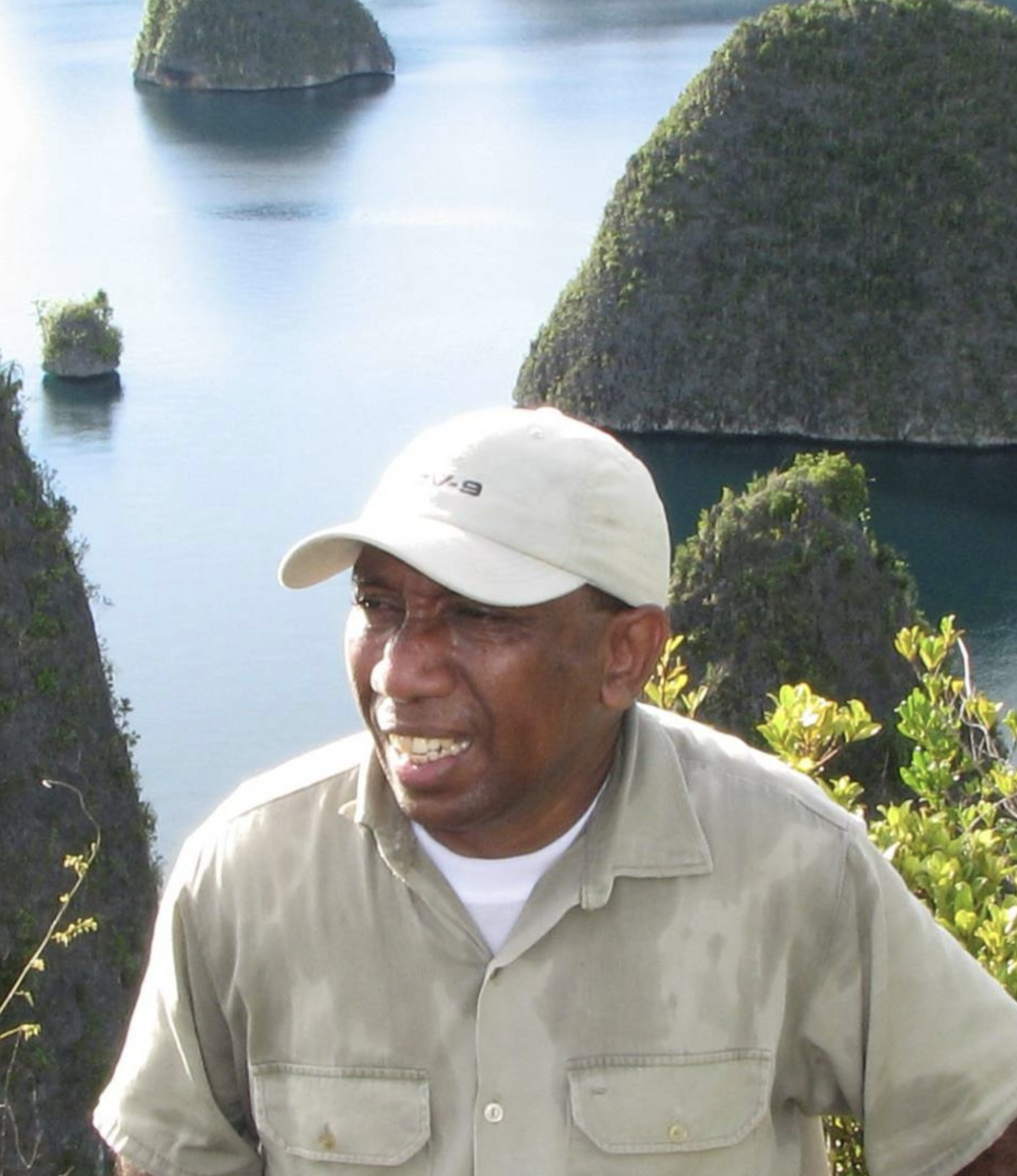 A man wearing a white cap and beige shirt standing outdoors with a scenic ocean and rocky islands in the background.