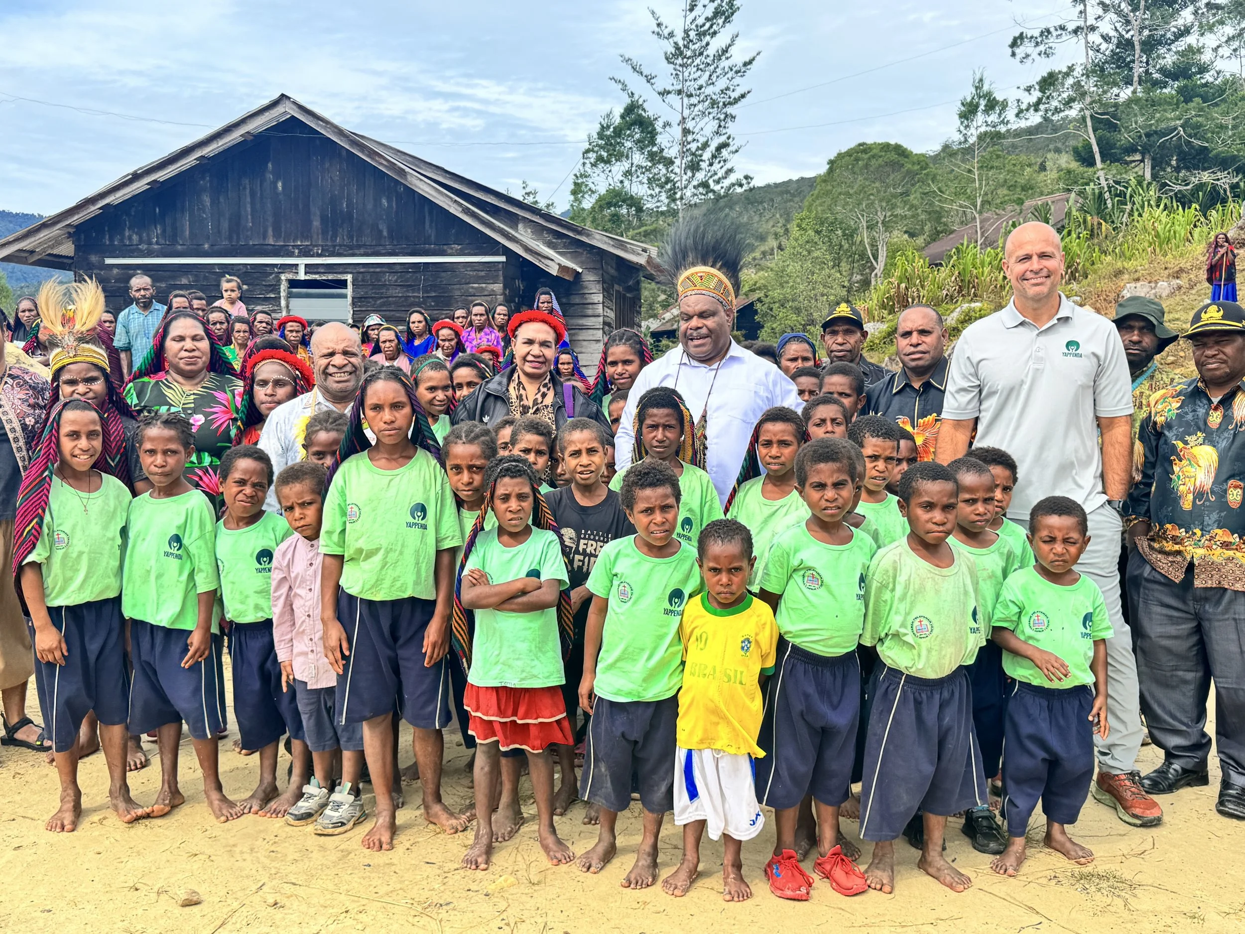 A large group of people, including children and adults, gathered outdoors in front of a wooden house, with some dressed in traditional indigenous clothing and others in casual attire, against a backdrop of trees and hills.