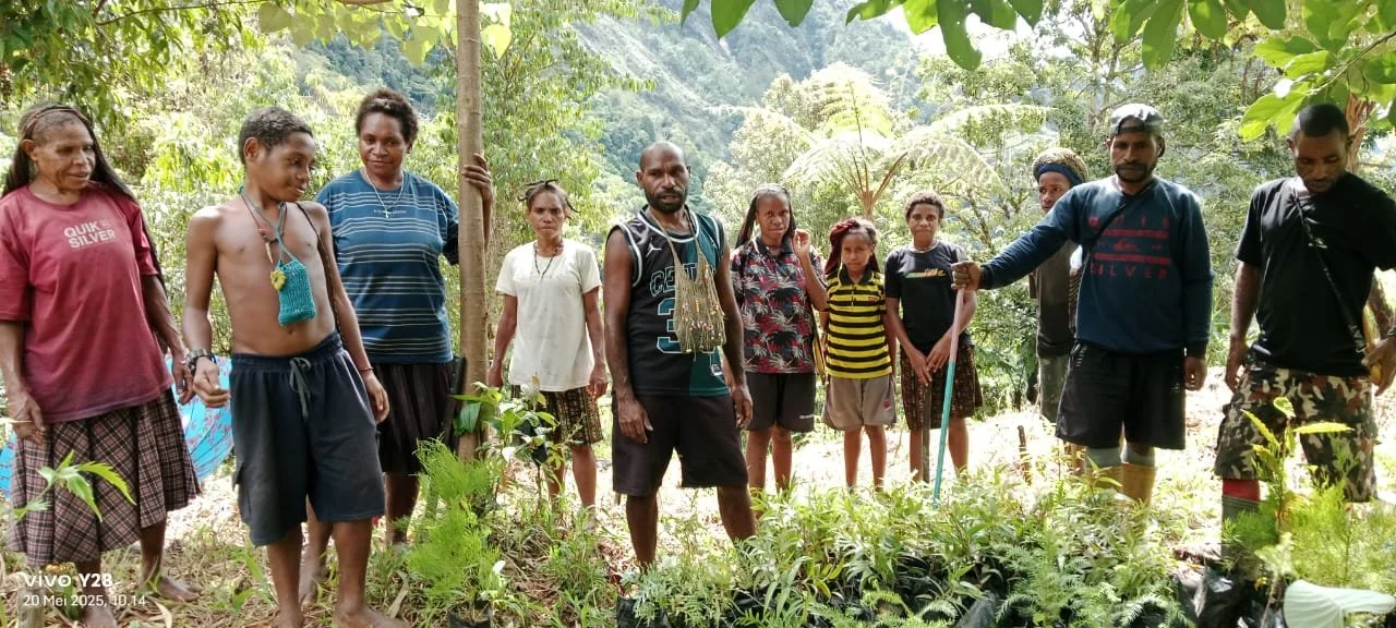 Group of people standing outdoors in a lush, green forest area, some children and adults, with a mountain in the background, participating in a community activity involving plants.
