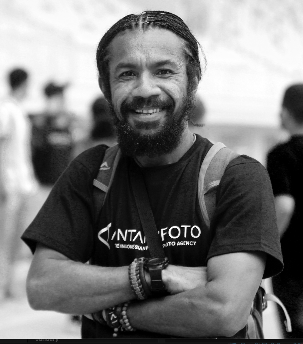 A smiling man with a beard and dreadlocks, wearing a black t-shirt and a backpack, standing outdoors with blurred people in the background.