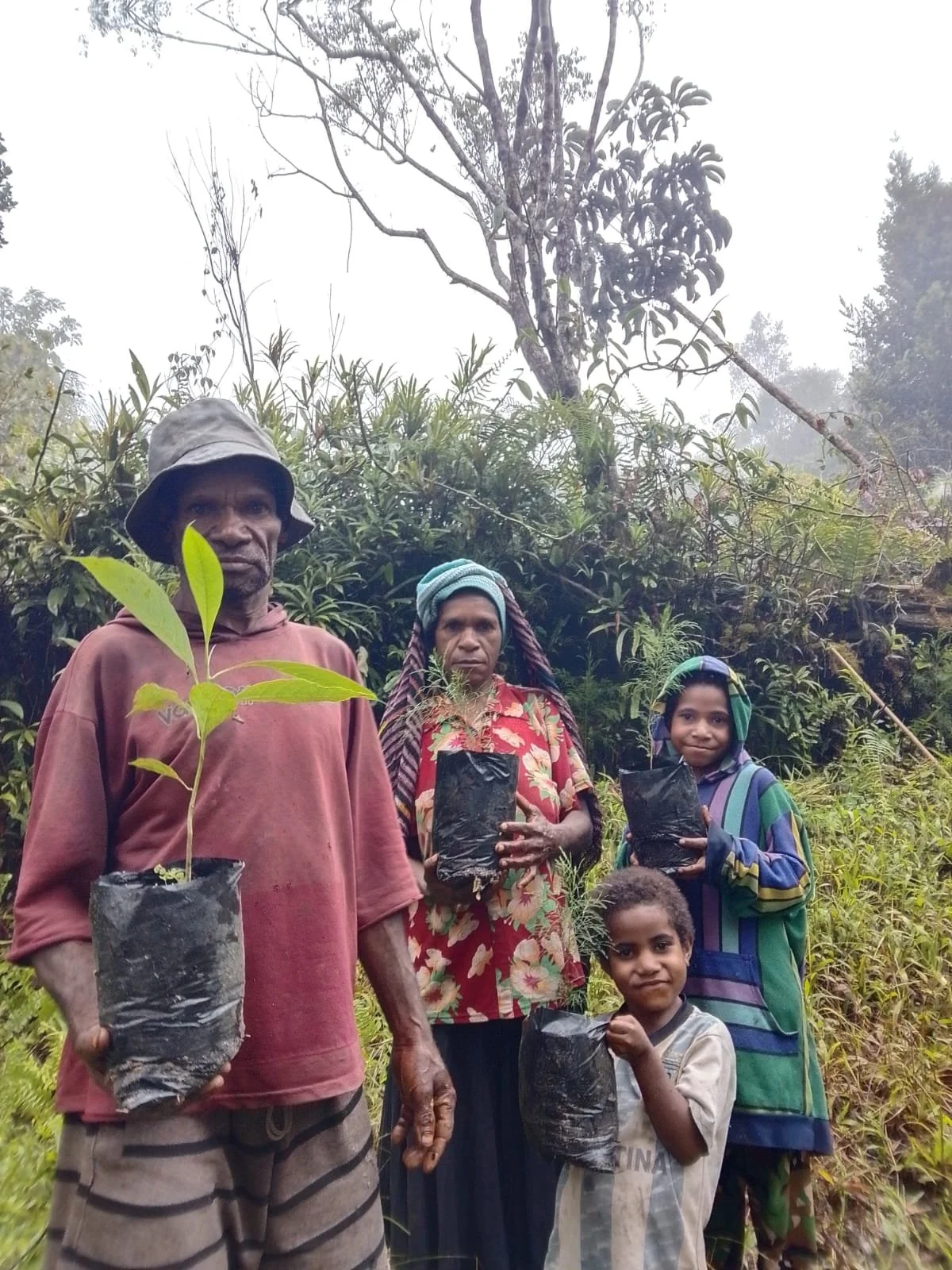 A group of five people, including three children and two adults, stand outdoors in a lush, green area holding young tree saplings in black plastic bags, ready for planting.