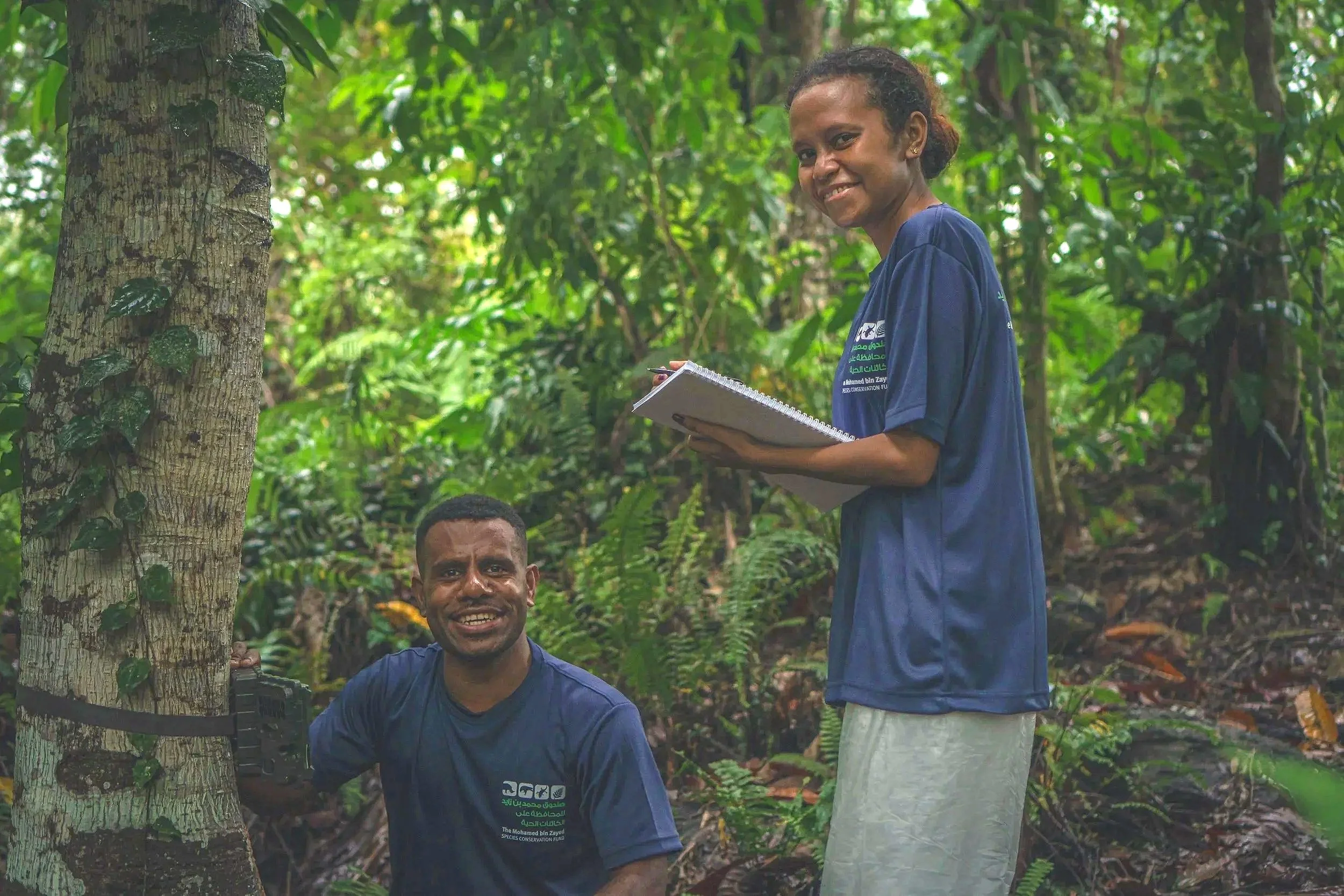 Two men and a woman are in a lush green forest, with one man beside a tree, another woman standing and holding a notebook, and a man sitting and smiling.