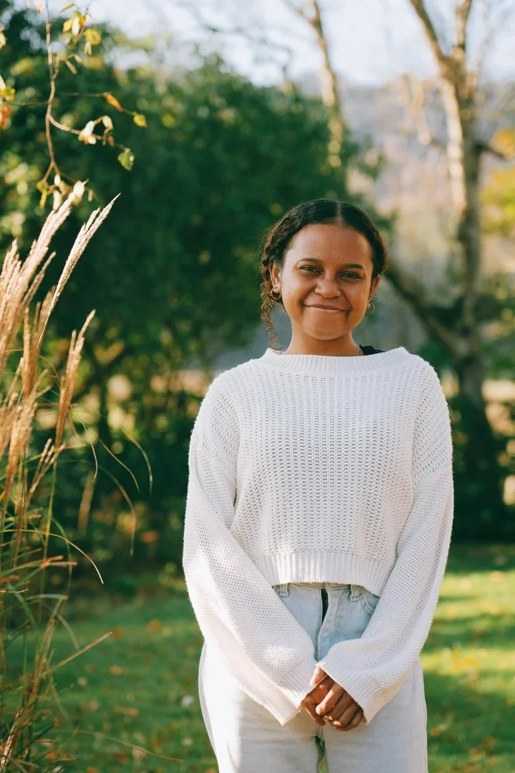 Smiling woman in a white sweater and light jeans standing outdoors in a park with trees and greenery in the background.