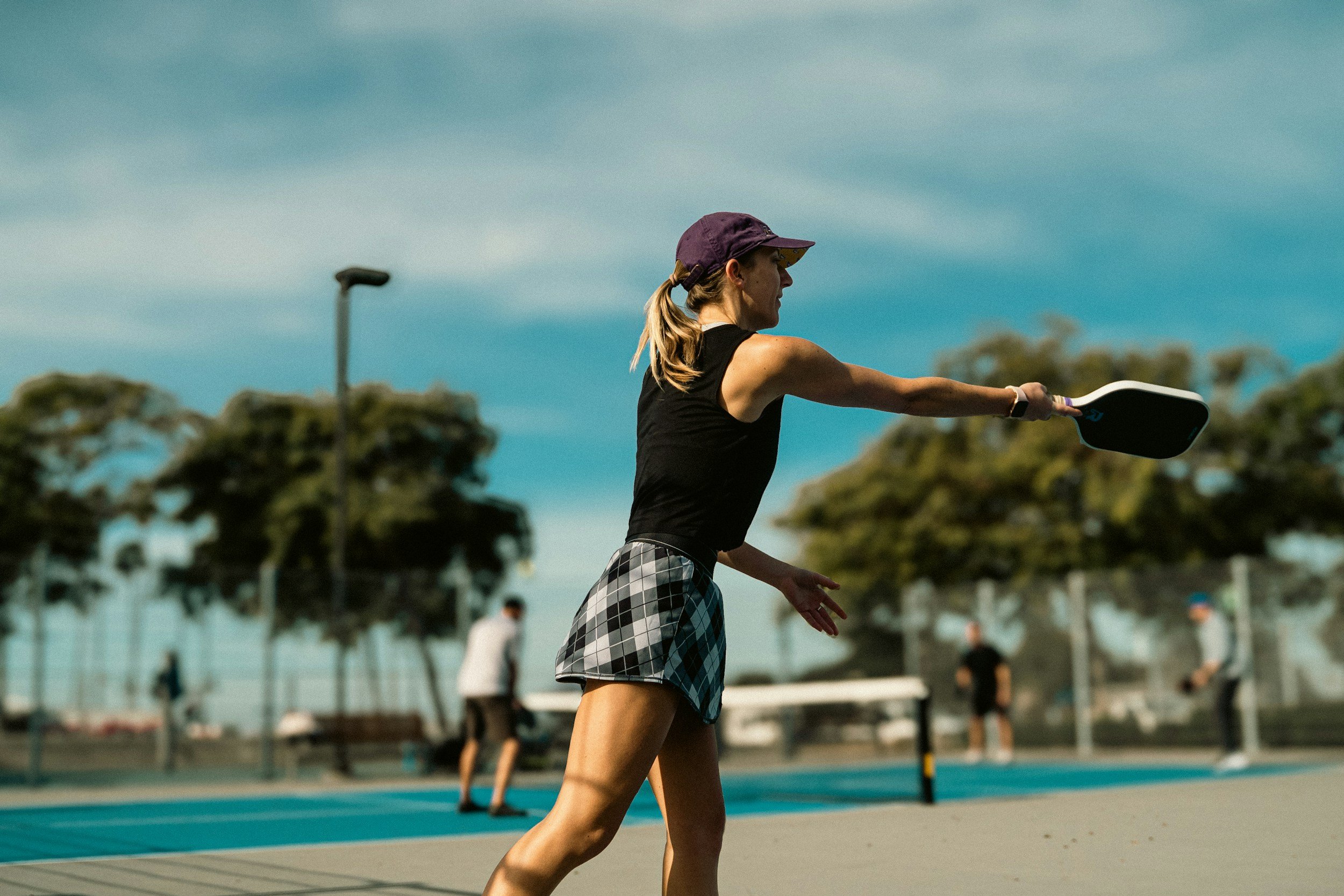 A woman playing pickleball on an outdoor court, wearing a purple cap, black tank top, and plaid shorts, with other players and trees in the background.