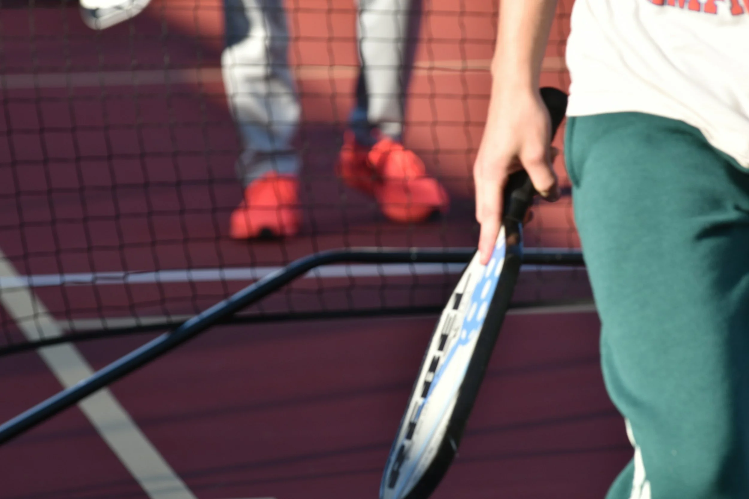 Close-up of a person's hand holding a tennis racket on a tennis court with a red wall and tennis net in the background.