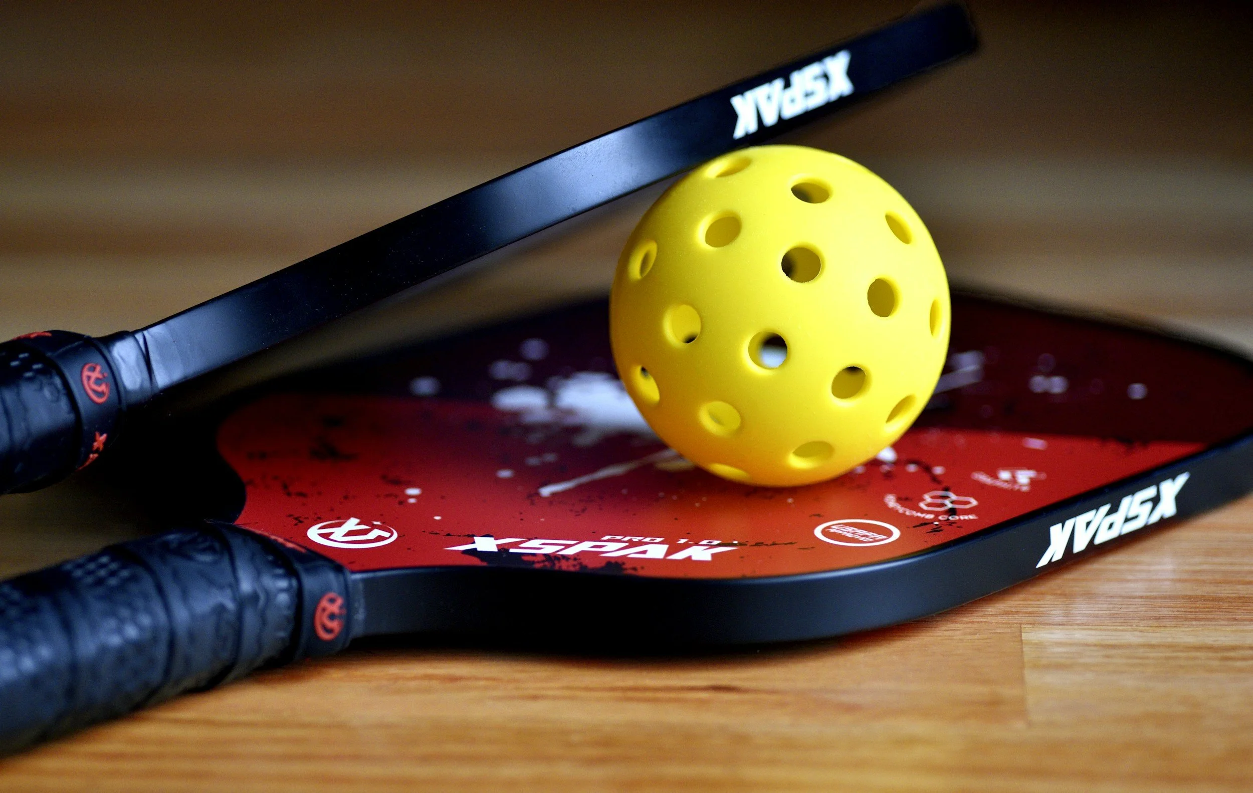 A YS Play tennis racket with black handle grips, a yellow perforated rubber ball, and a red YS Play tennis mat on a wooden surface.