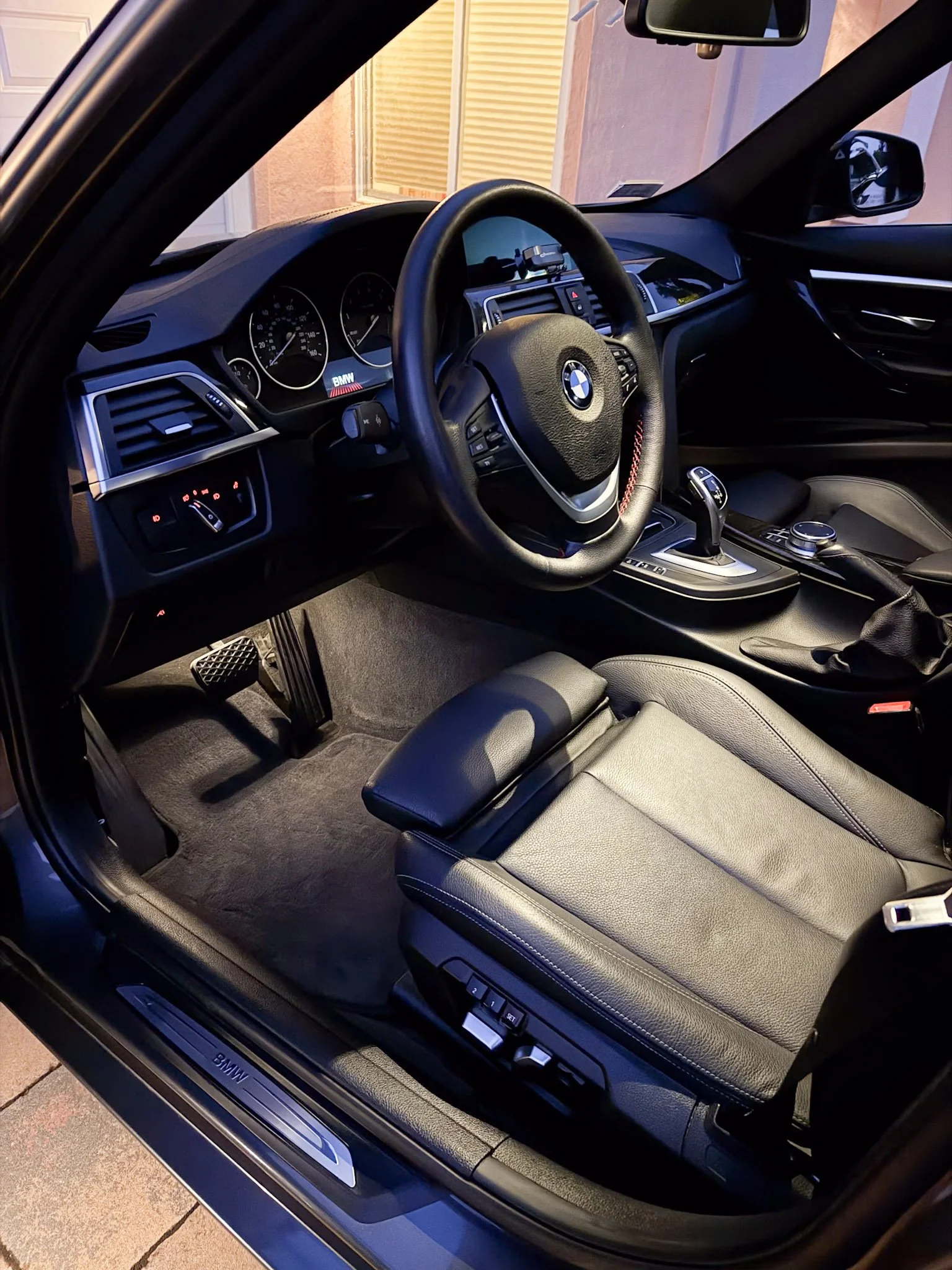 Inside view of a BMW car showing the steering wheel, dashboard with gauges, gear shift, and front passenger seat, taken from the open driver's side door.