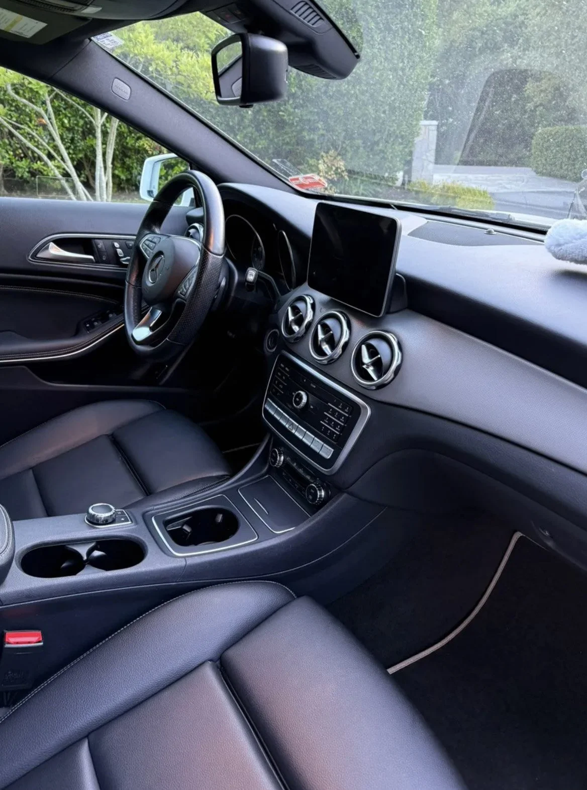 Interior of a modern car showing the dashboard, steering wheel, center console, and black leather seats, with a view of greenery through the windshield.