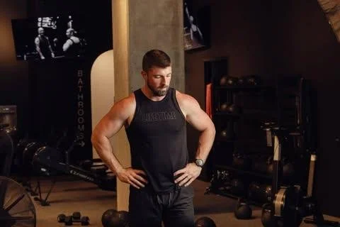 A man in a gym wearing a sleeveless black shirt and shorts, standing with his hands on his waist, looking down.