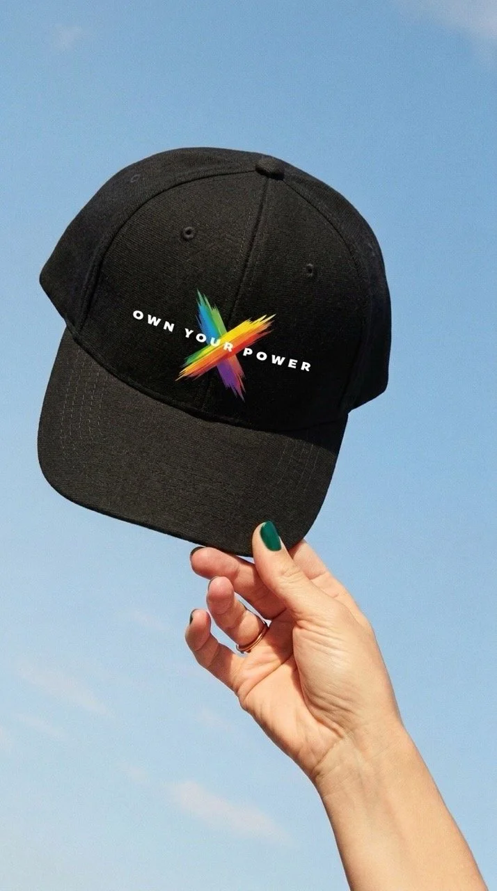 A hand holding a black Own your power baseball cap by Pickleball Merch Aus, against a clear blue sky.