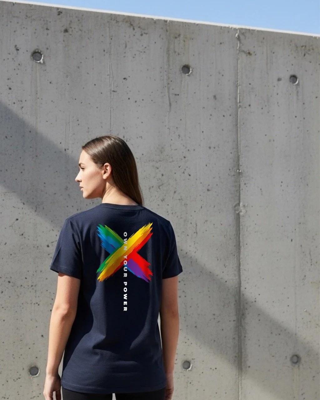 A model wearing a navy T-shirt by Pickleball Merch Aus, with a colourful rainbow 'X' and 'Own Your Power' text on the back. She is standing against a concrete wall.