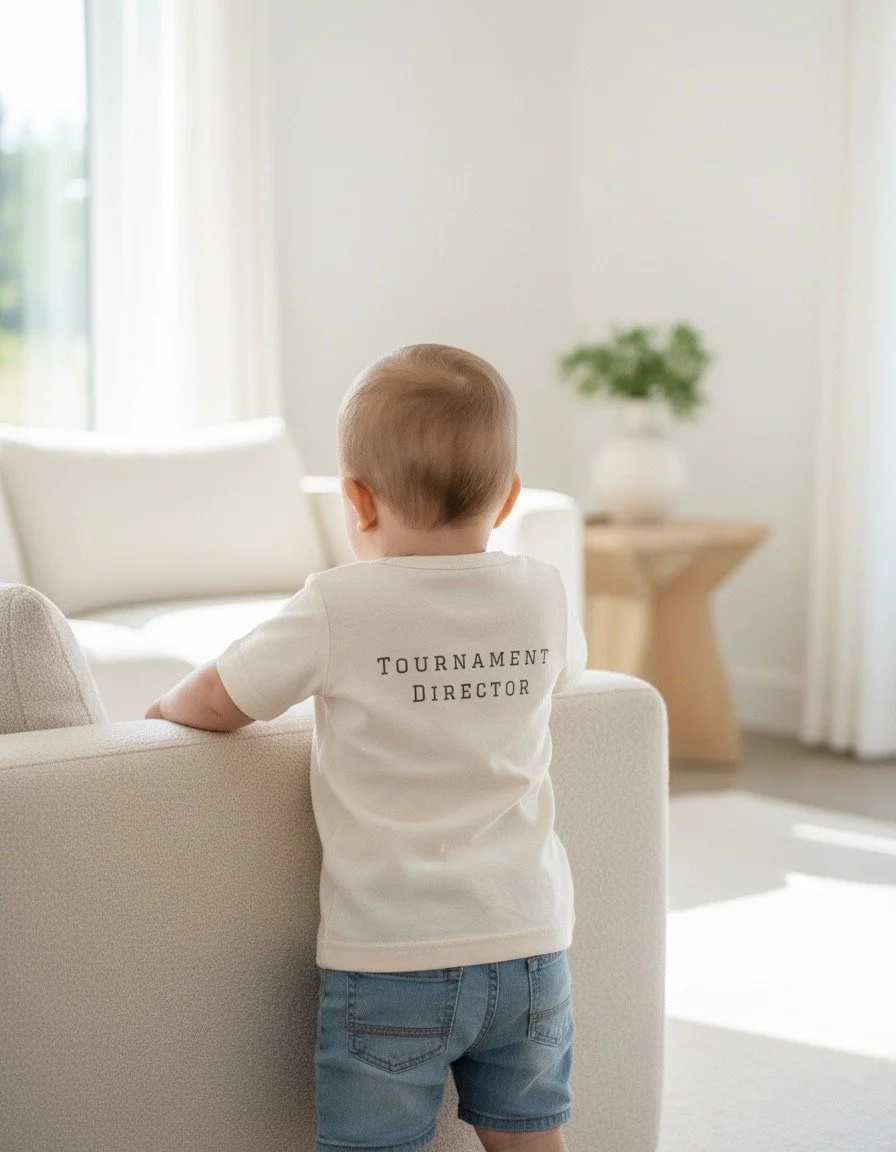 An infant wearing a cream pickleball wee tee by Pickleball Merch Aus, with the words Tournament Director on the back, standing in a playroom.