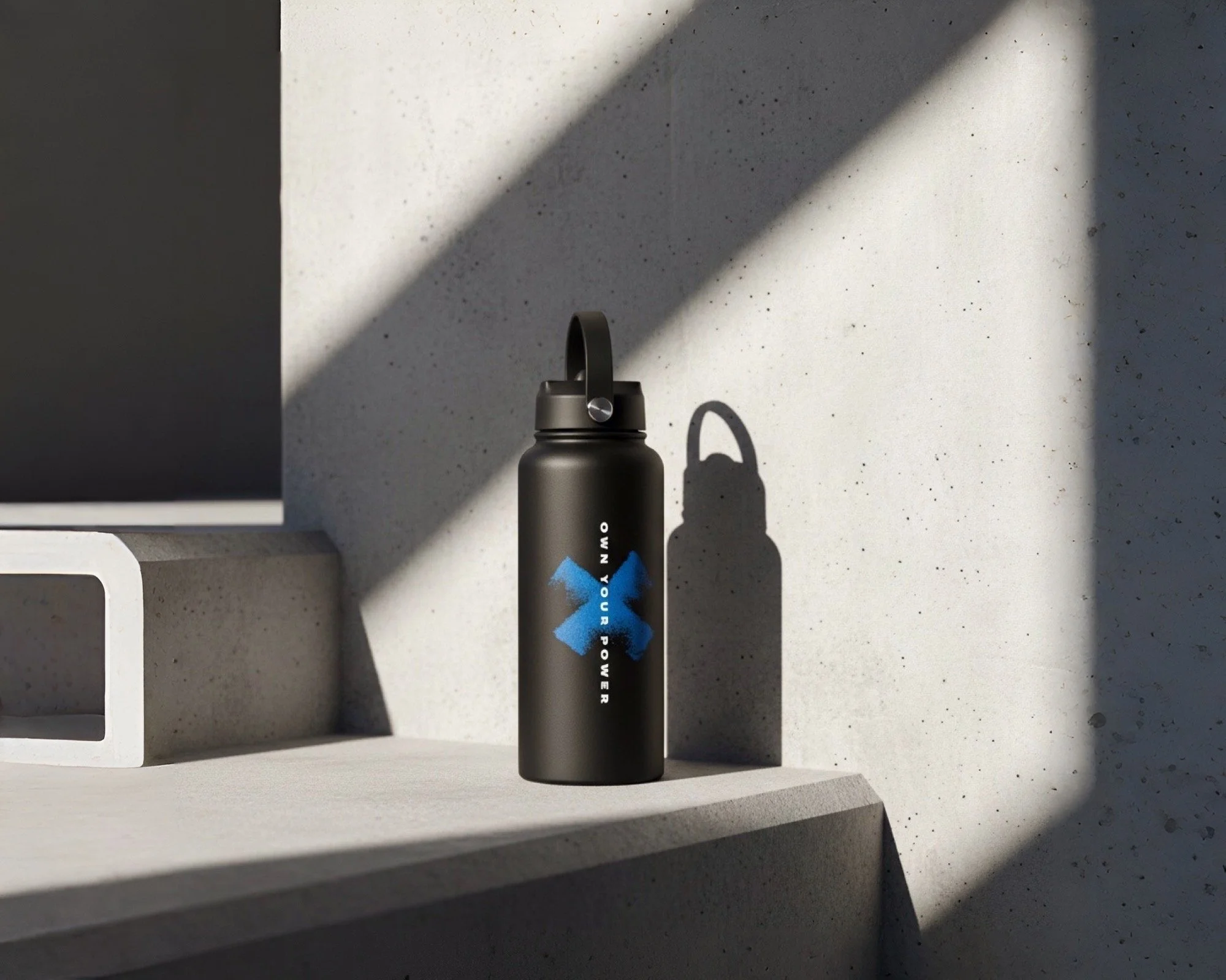 A black water bottle with a blue logo and silver cap standing on a concrete surface with a shadow cast on a white wall behind it.