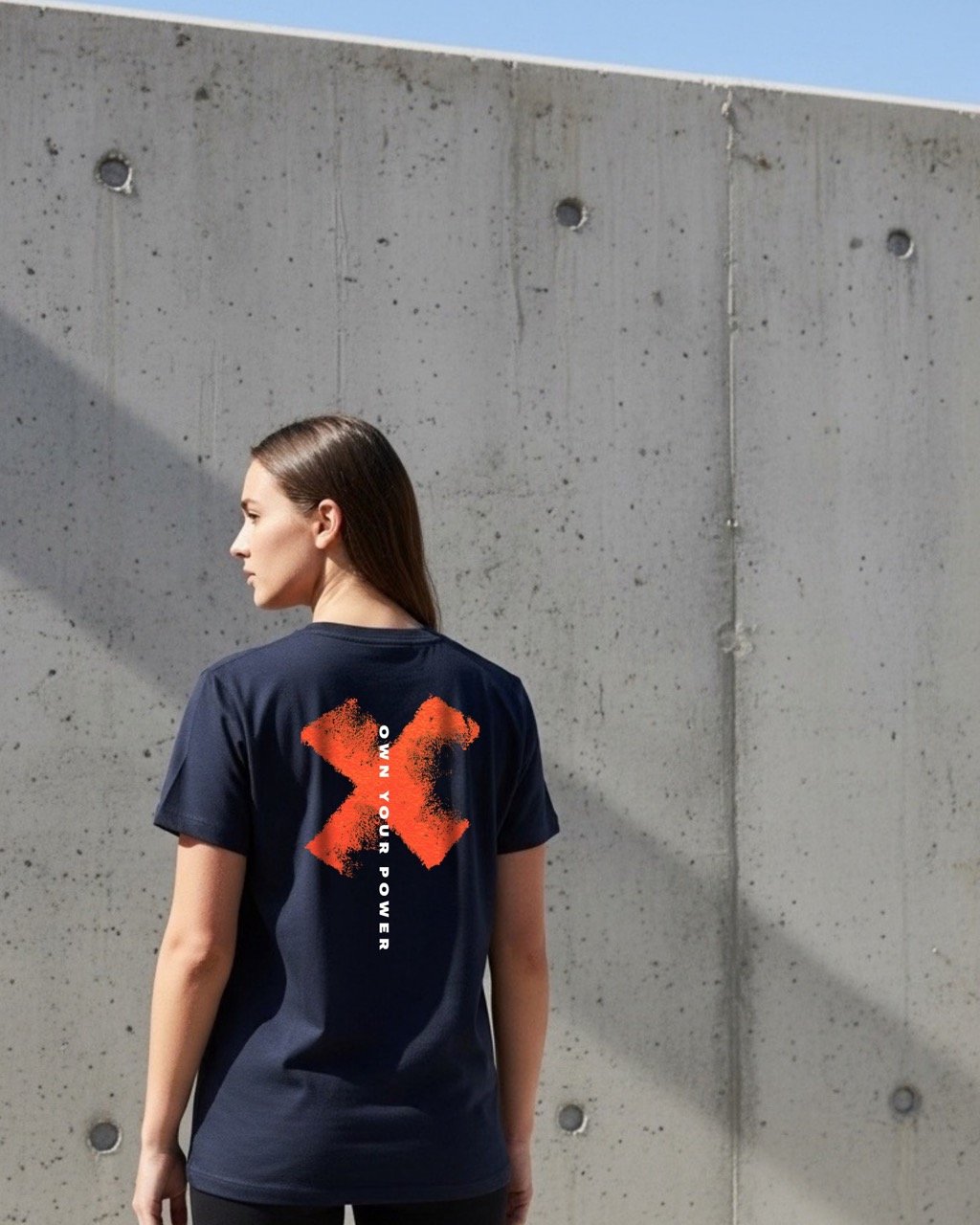 A woman with long brown hair standing outdoors against a concrete wall, wearing a navy blue t-shirt with red and white graphic and text on the back that reads 'OWN YOUR POWER'.