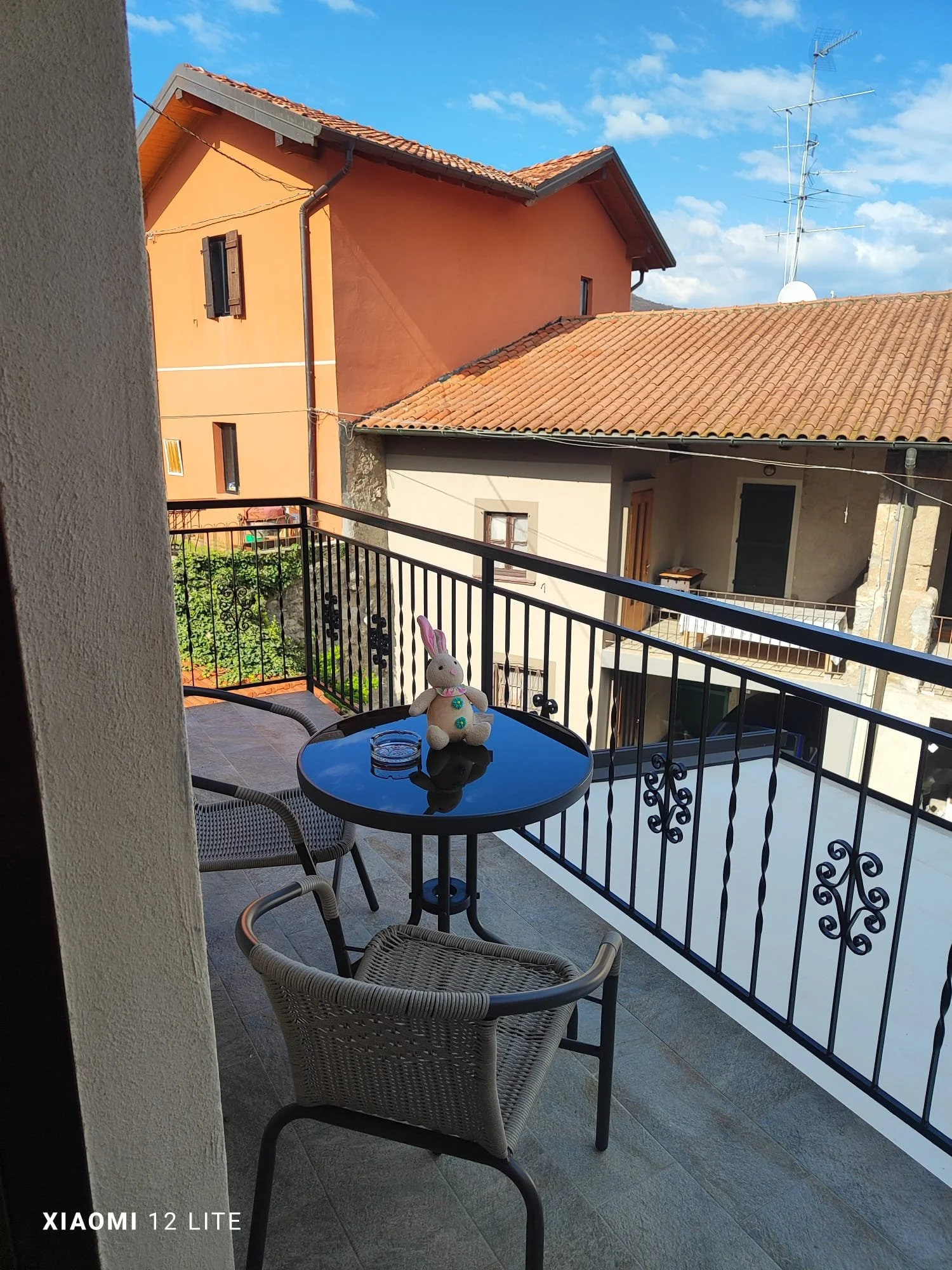 Balcone con tavolino, due sedie, orsetto di peluche e due bicchieri, con vista su case con tetti di tegole e antenne, sotto un cielo blu con nuvole.